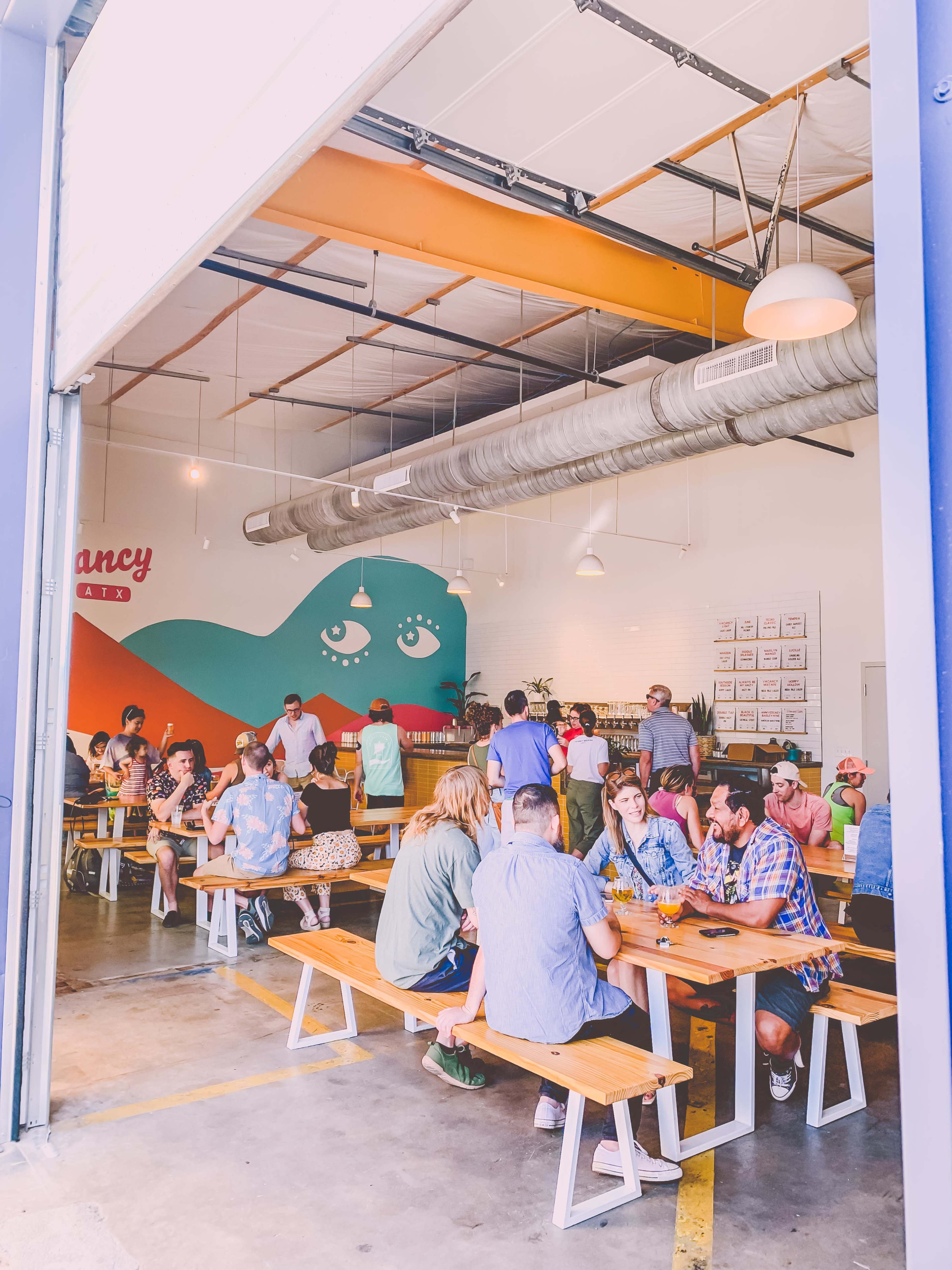 A vibrant casual dining space features colorful murals and groups of people seated at wooden benches enjoying food and drinks.