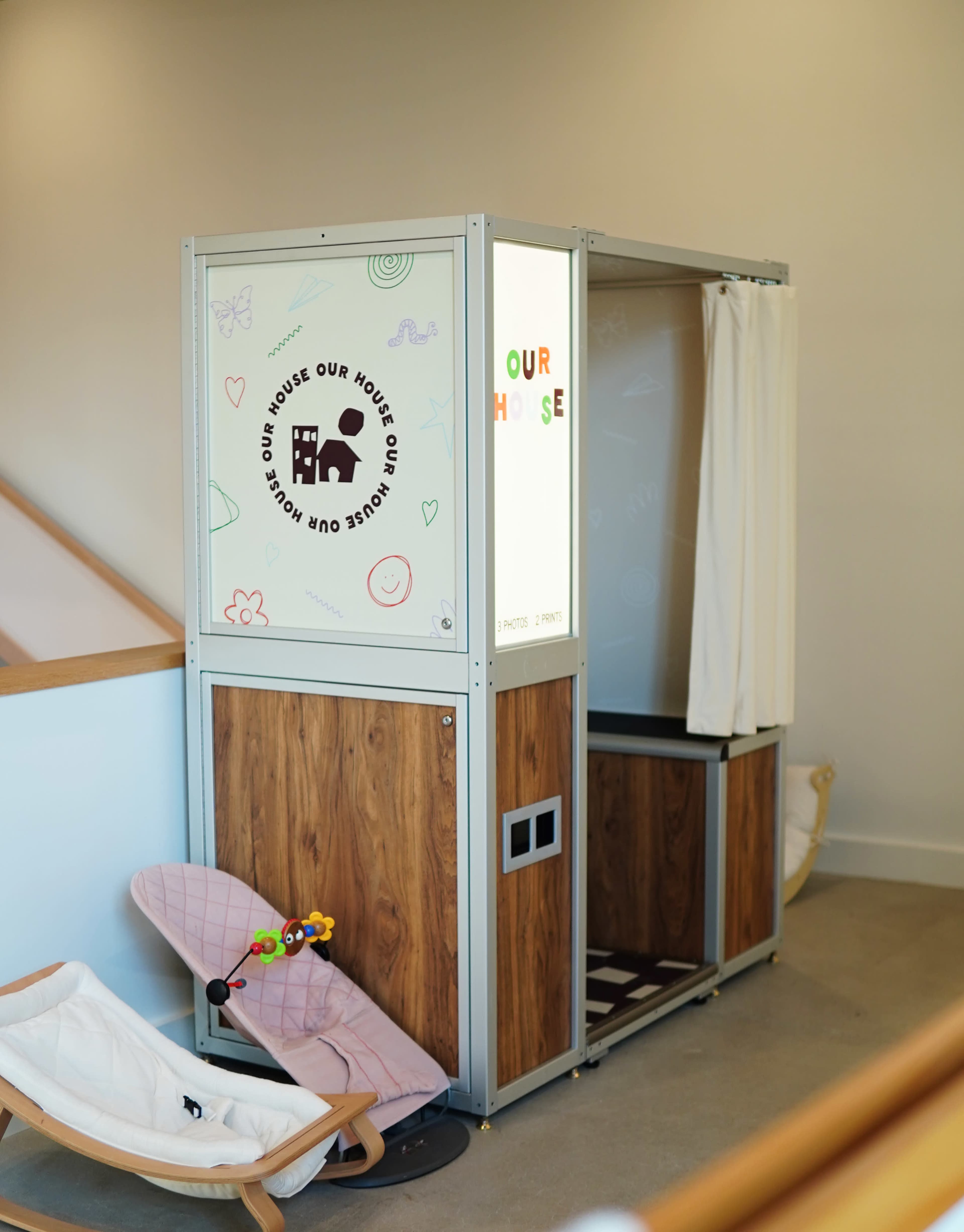 The image shows a colorful play booth with wooden accents and a light-up sign that reads "OUR HOUSE," situated in a well-lit indoor space.