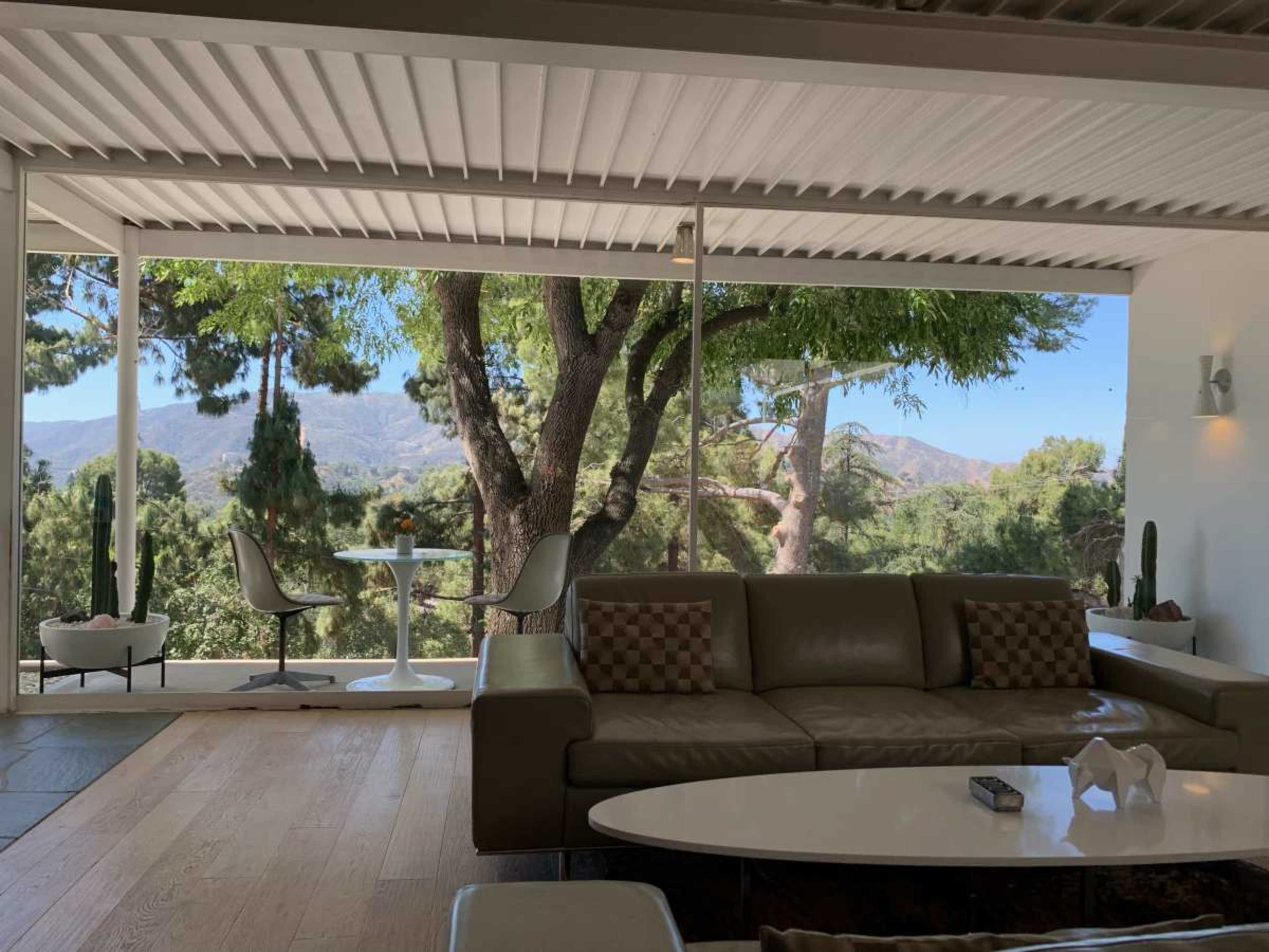 The image shows a modern interior living space with a sofa and coffee table, overlooking a scenic view of mountains and trees through large windows.