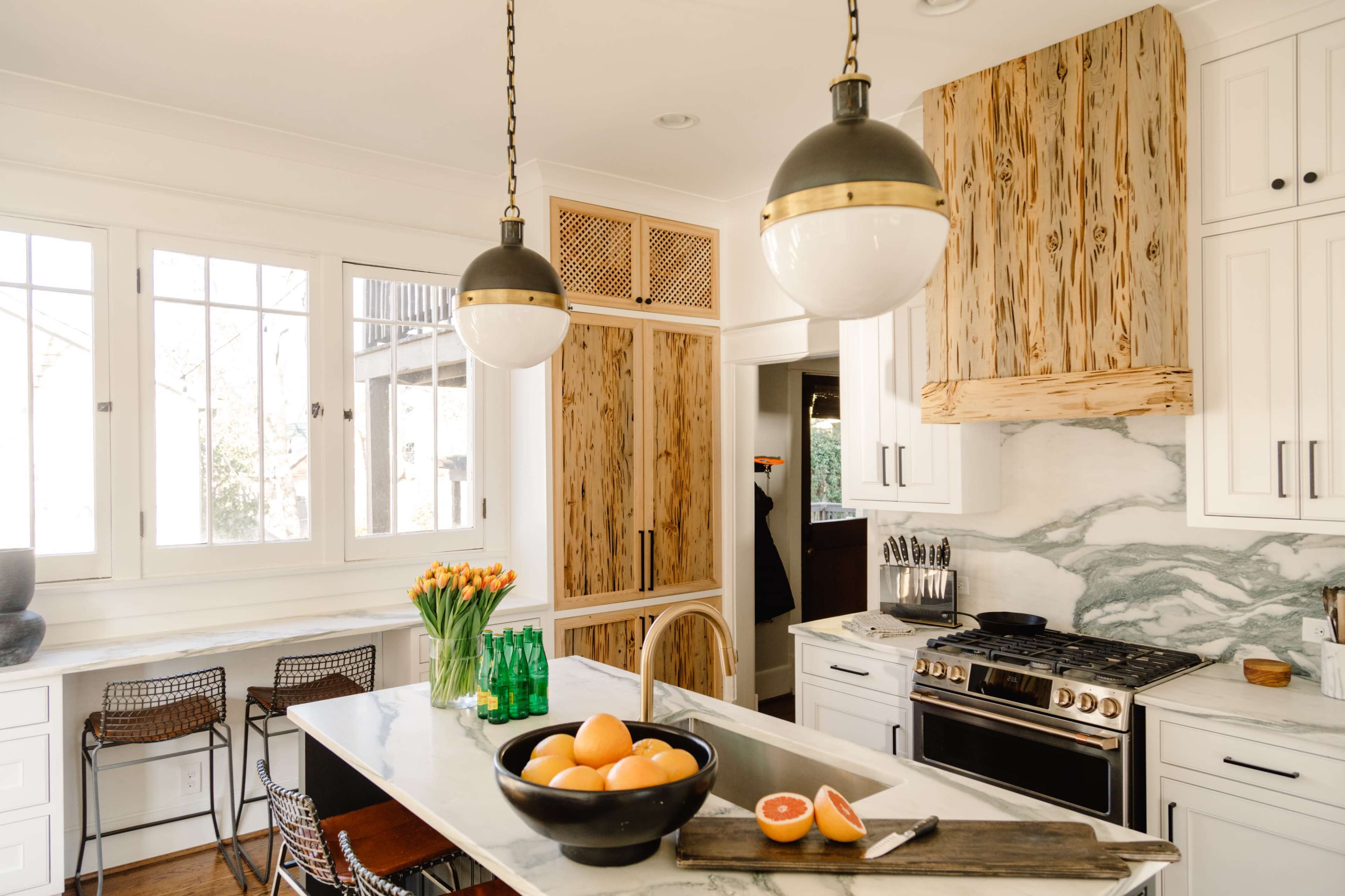 A modern kitchen features a marble island with a black bowl of oranges, wooden cabinetry, and pendant lights.