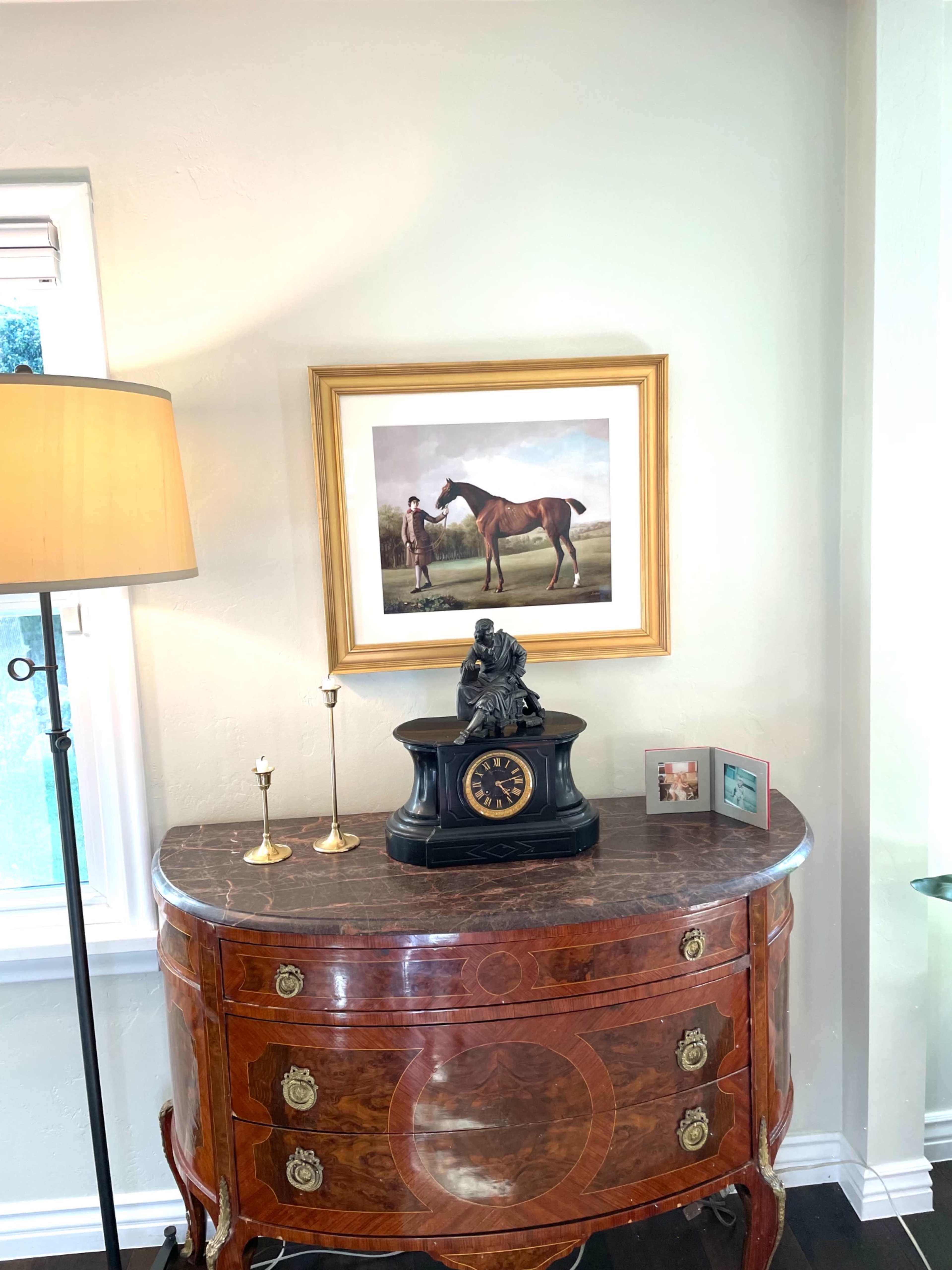 A wooden sideboard with a clock sits beneath a framed painting of a horse and rider, accompanied by a lamp and two small photo frames.