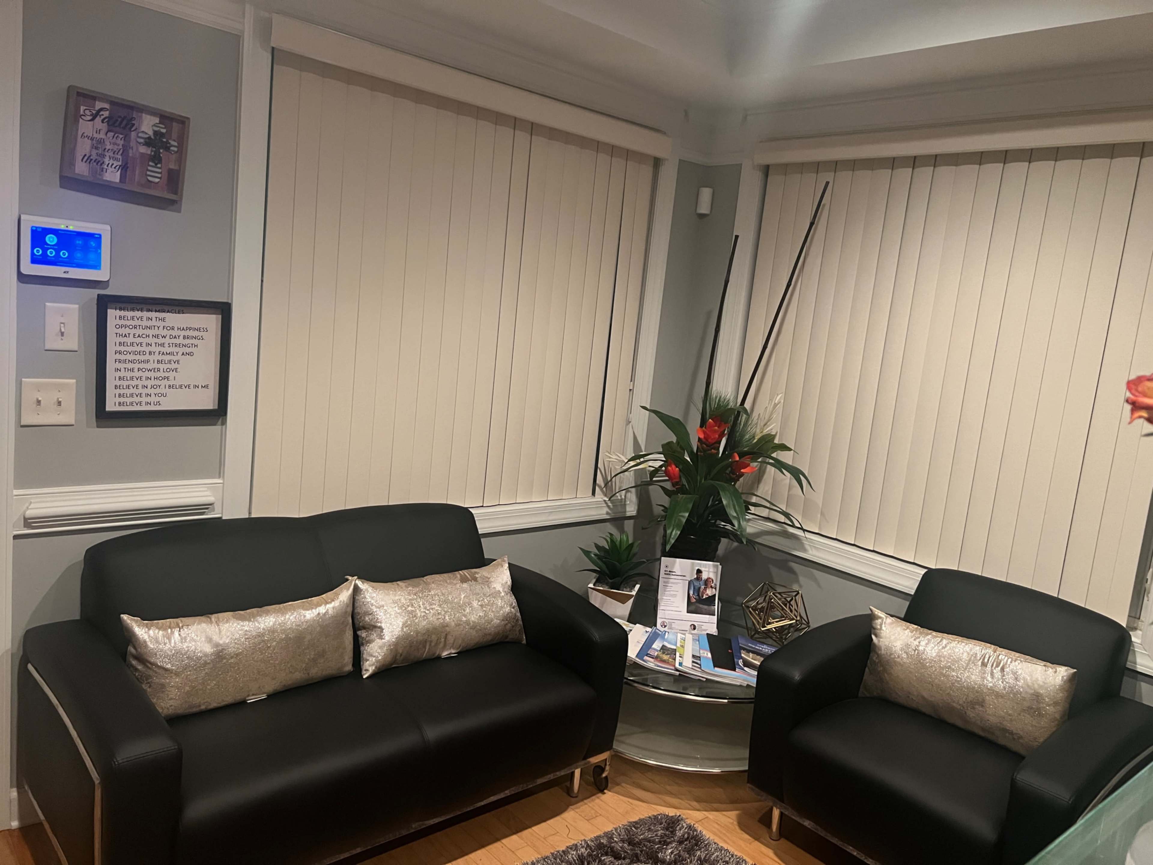 The image shows a cozy waiting area with a black sofa and armchair, a glass coffee table, and potted plants, set against light-colored walls and window blinds.