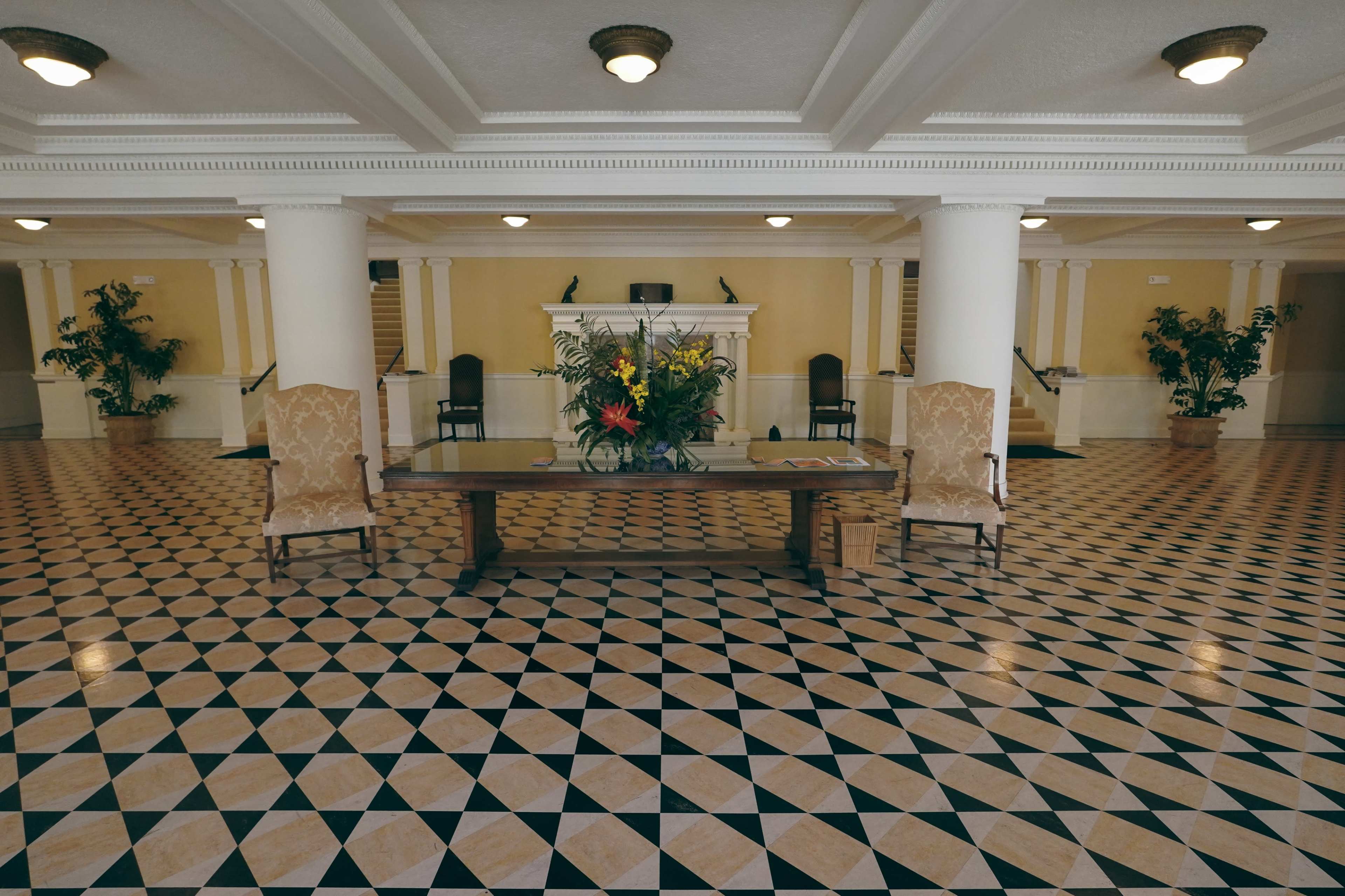 A spacious lobby featuring a patterned tile floor, a central wooden table with a flower arrangement, and decorative armchairs beside two large pillars.