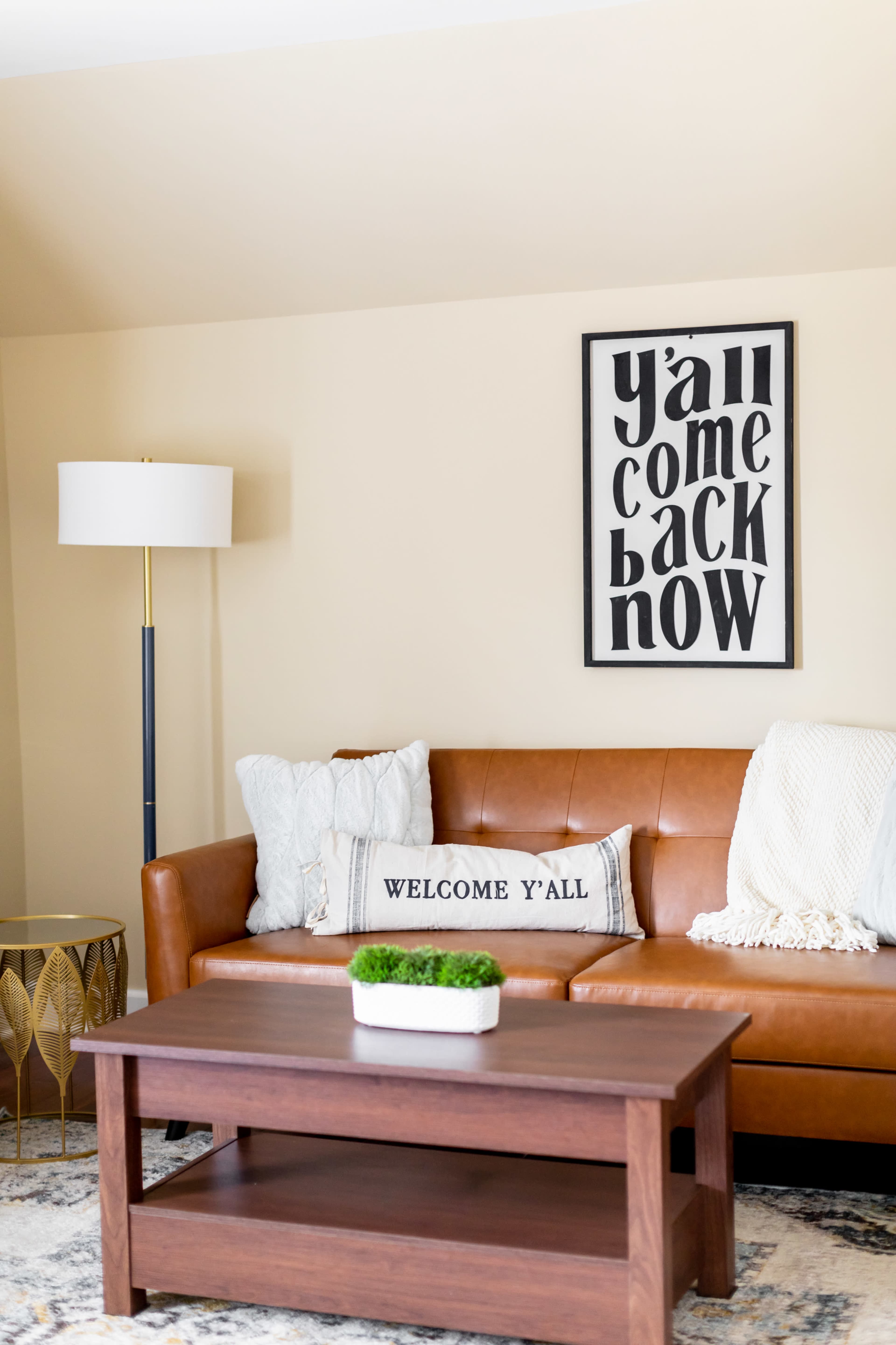A cozy living room with a brown leather sofa, decorative pillows, a wooden coffee table, and a wall art piece that reads "y'all come back now."