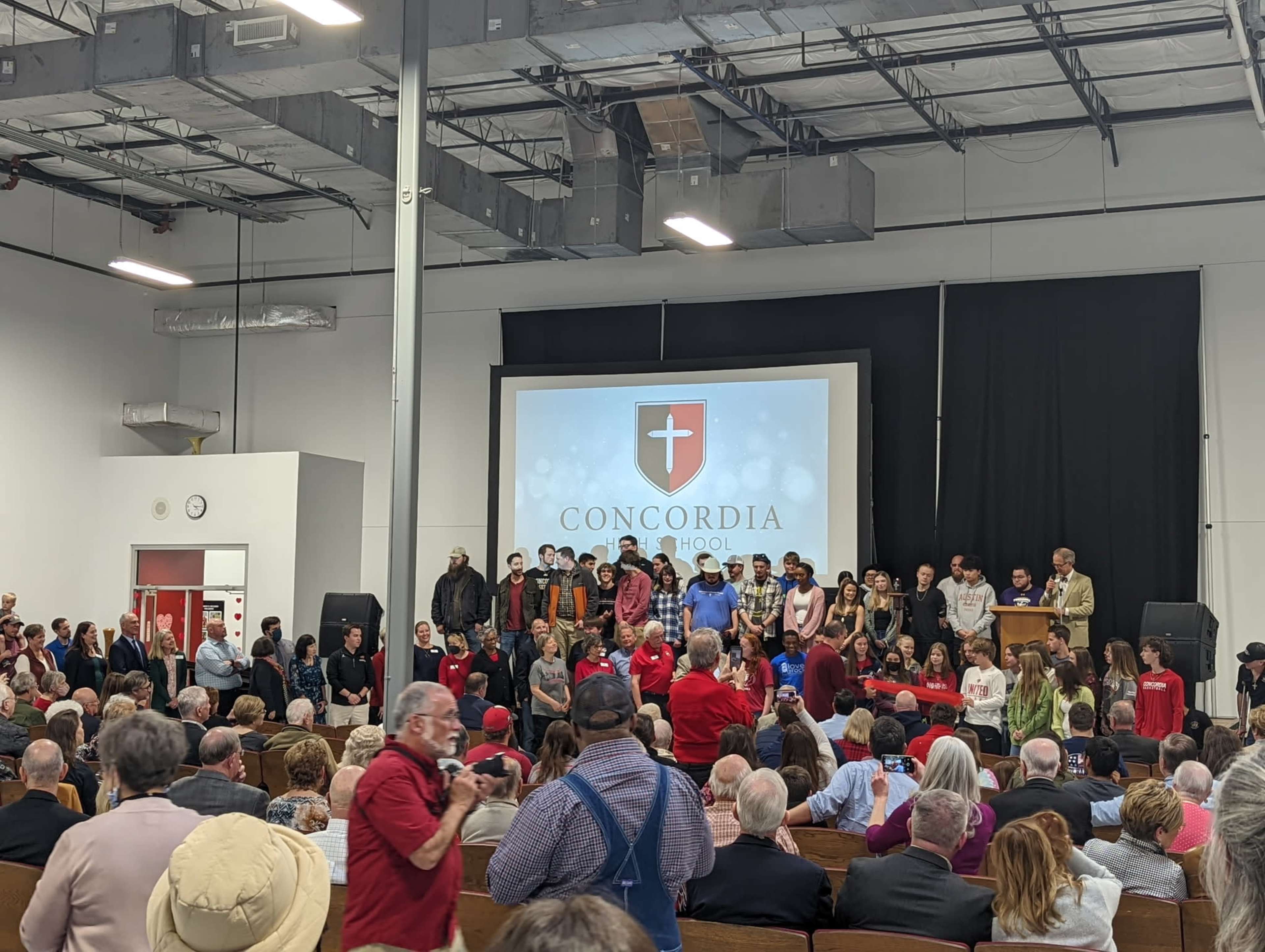 A large group of people stands on a stage in front of an audience, with a screen displaying the logo of Concordia School in the background.