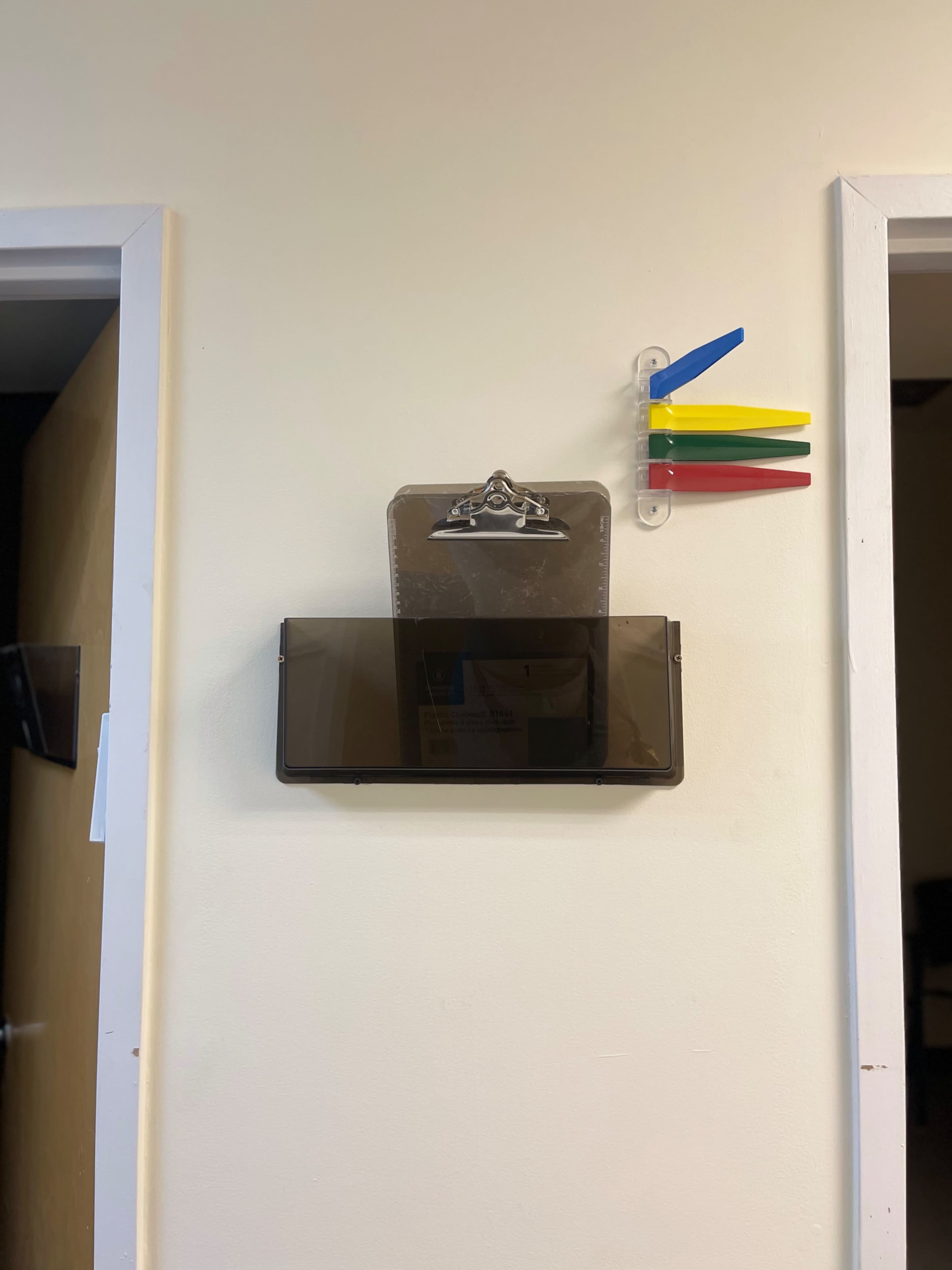 A clipboard holder with a transparent clip is mounted on a wall between two doors, accompanied by colorful plastic markers.