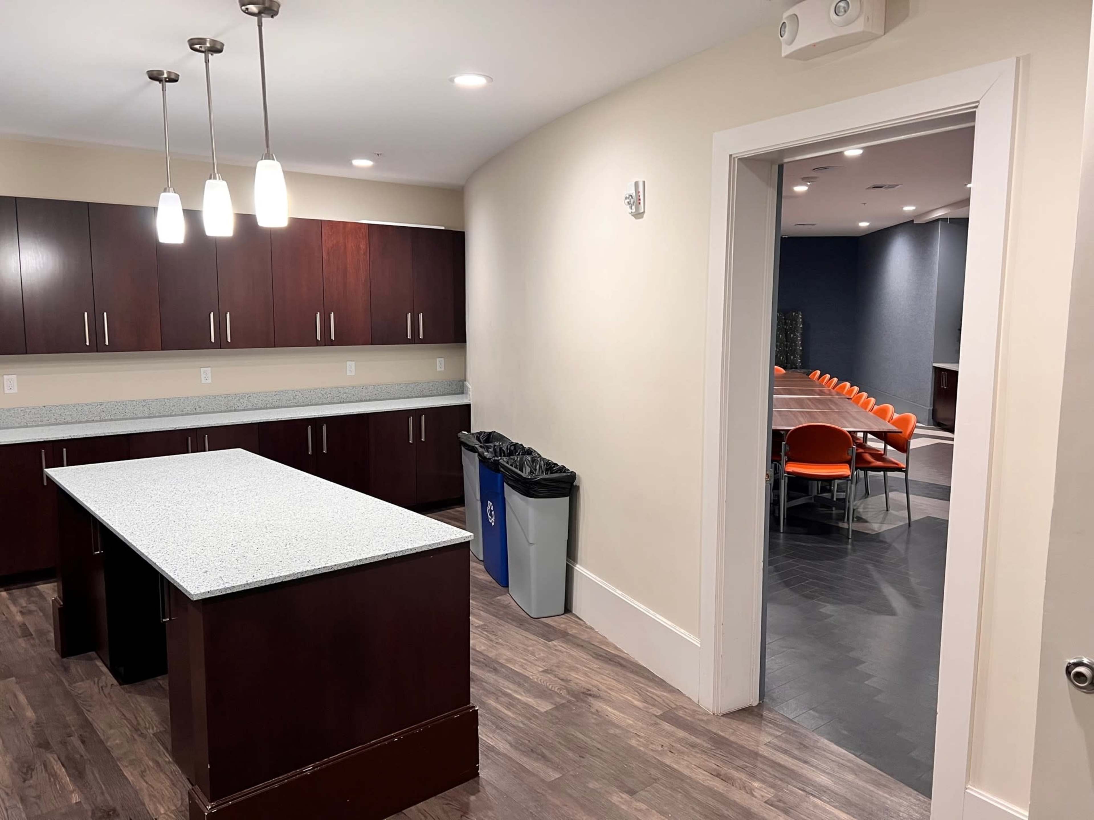 A modern kitchen area with a granite countertop island, dark cabinetry, and an open doorway leading to a meeting room with orange chairs.