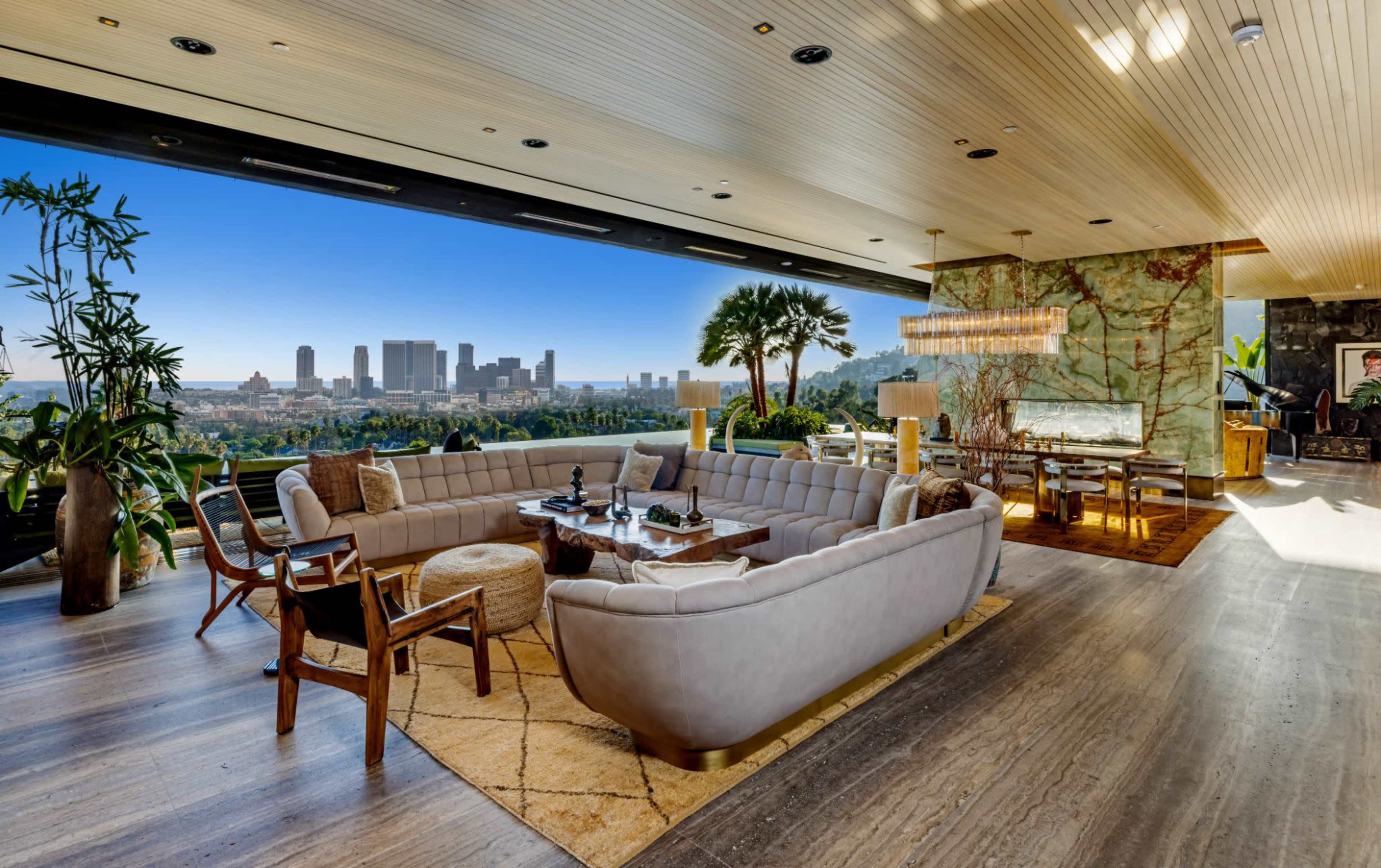 The image shows a modern living room featuring a large sectional sofa, a coffee table, and panoramic windows displaying a skyline view of a city.