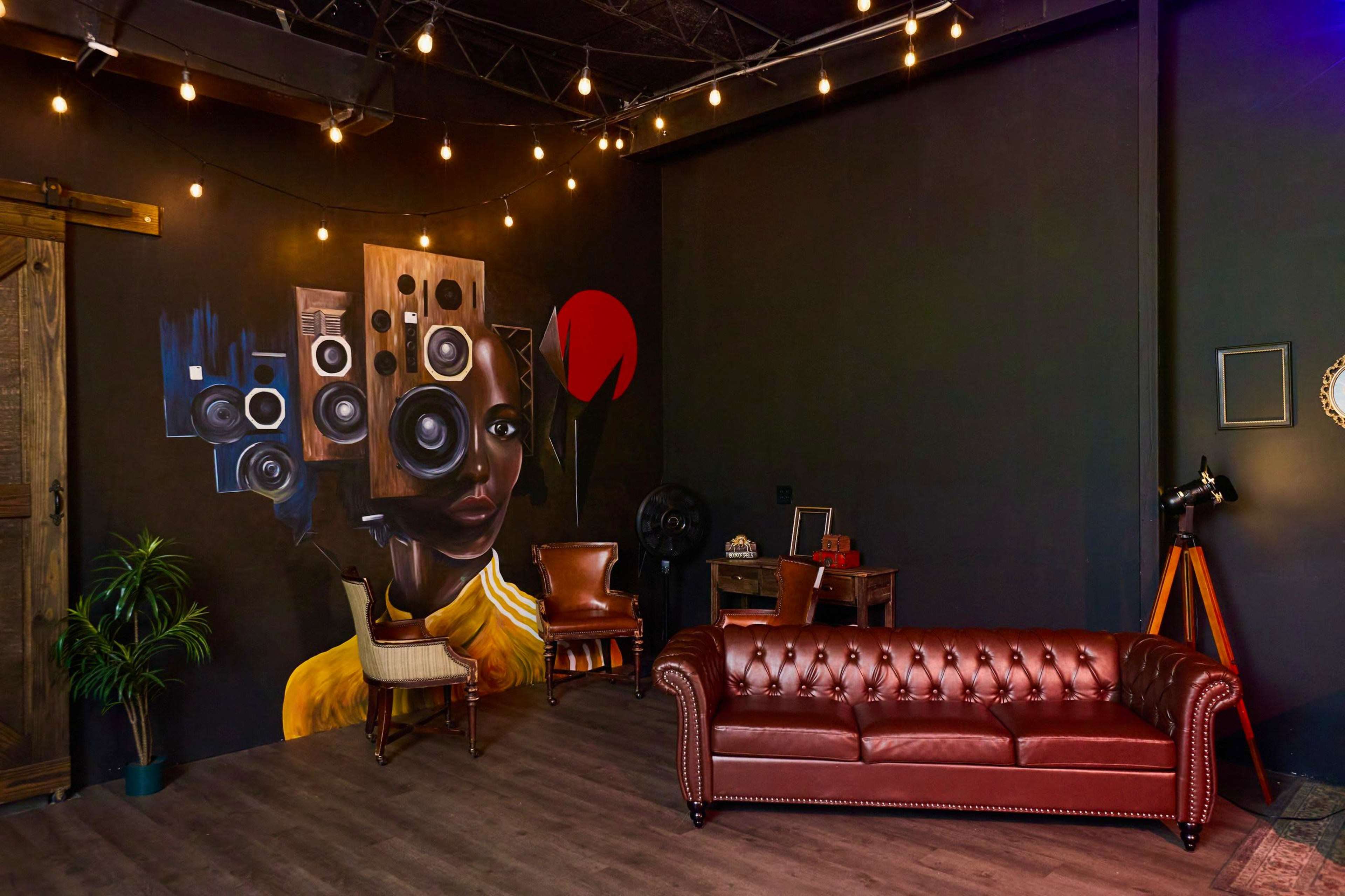 The image displays a cozy interior featuring a brown leather chesterfield sofa, wooden chairs, and a vibrant mural of a woman with speakers as a headdress, all under string lights.