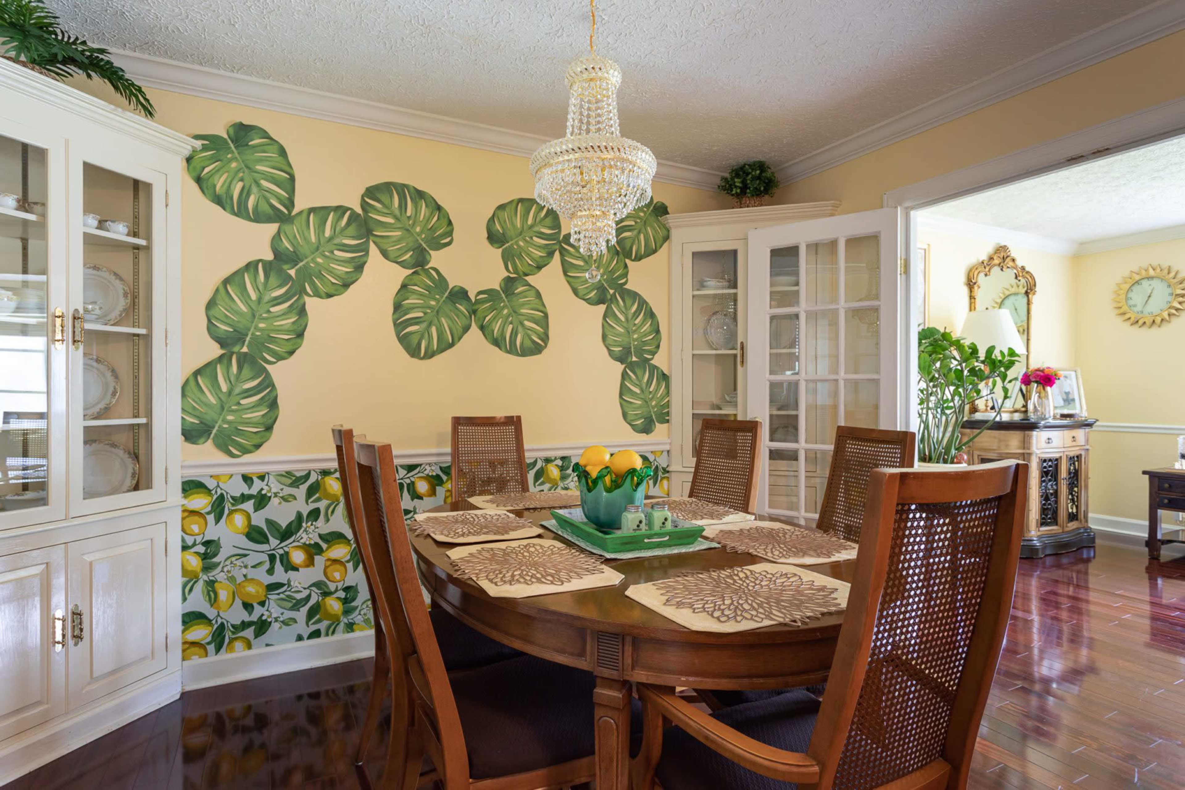 The dining room features a wooden table surrounded by chairs, with a chandelier above and a decorative mural of large leaves on the wall.