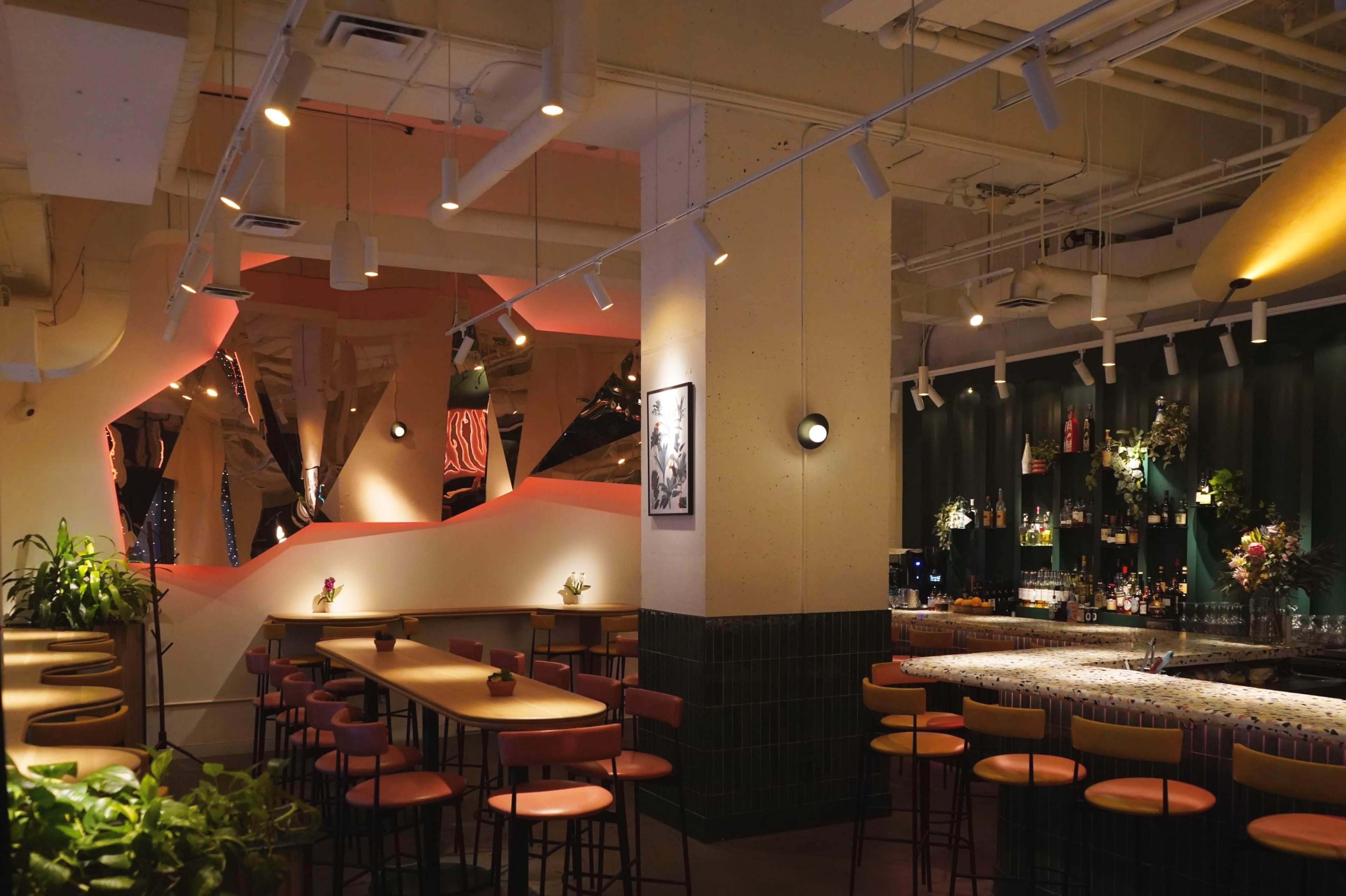 The image shows a modern restaurant interior featuring a bar area with an array of seating, decorative plants, and artistic wall designs.
