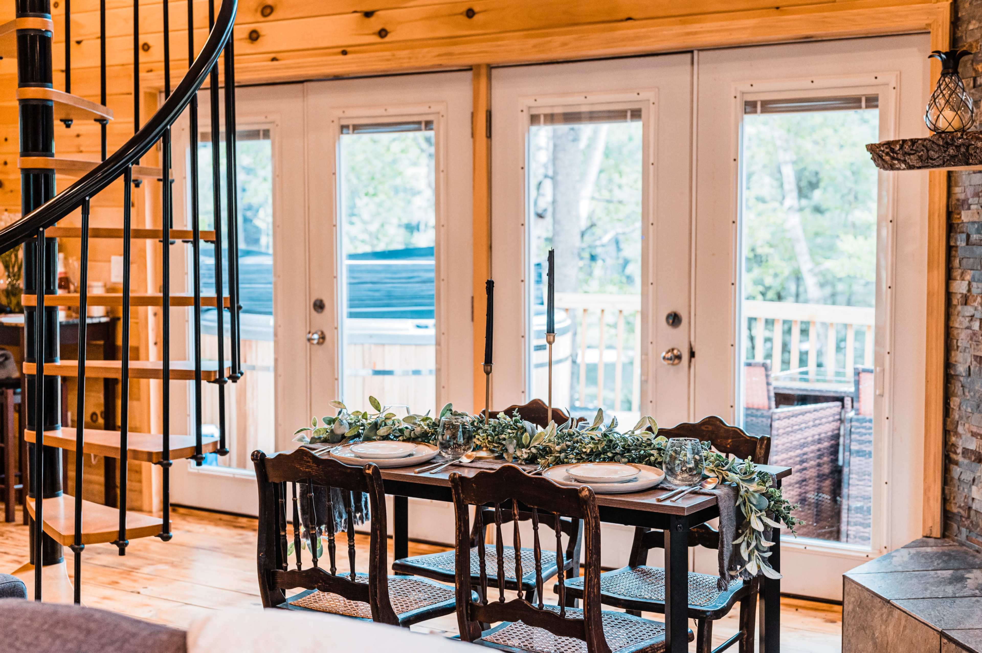 A wooden dining table set for a meal, positioned near large windows that open to a balcony with views of greenery.