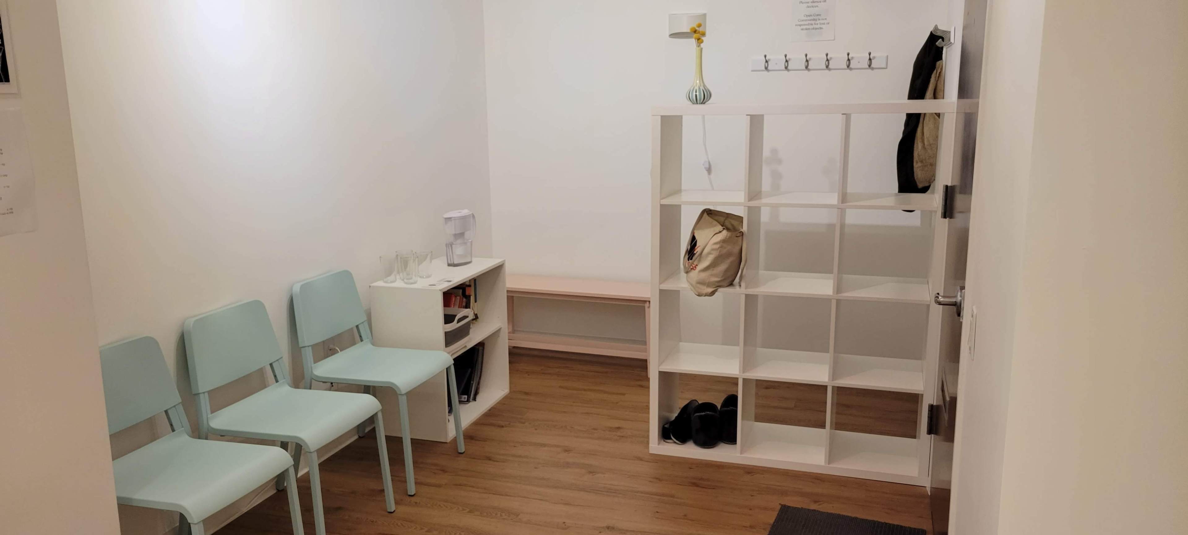 A small waiting area with two light blue chairs, a white shelf, a table, and a coat rack on a plain wall.