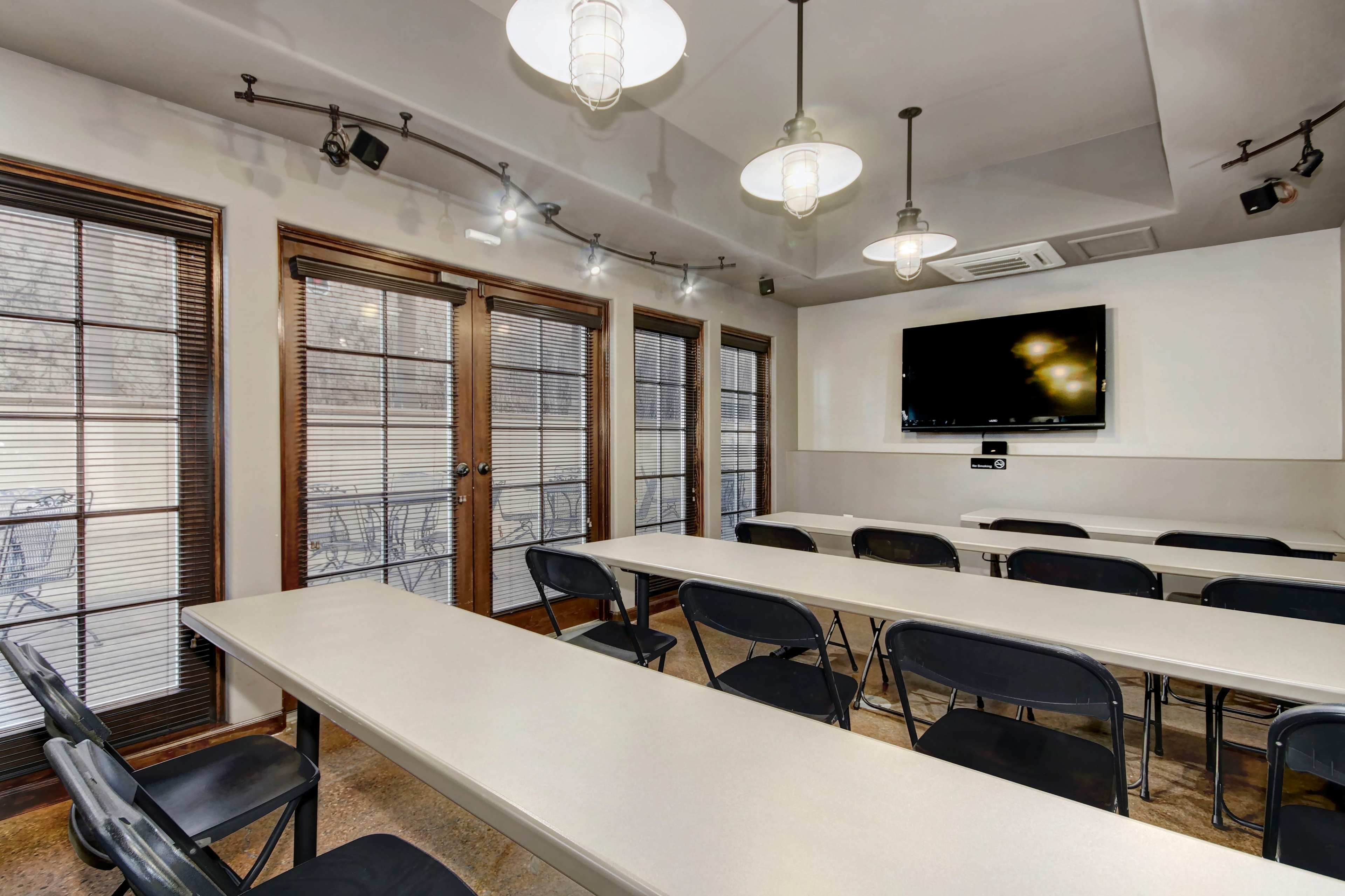 A meeting room features multiple tables and chairs arranged facing a wall-mounted television, with large windows providing natural light.