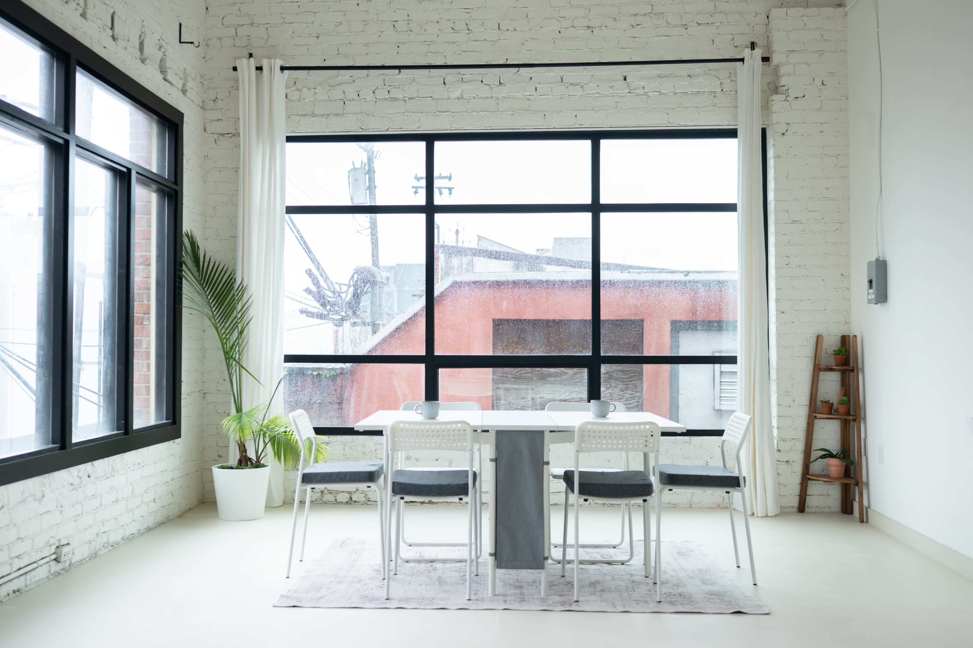 A modern dining area features a rectangular table surrounded by white chairs, positioned in front of large windows with a view of a rainy cityscape.