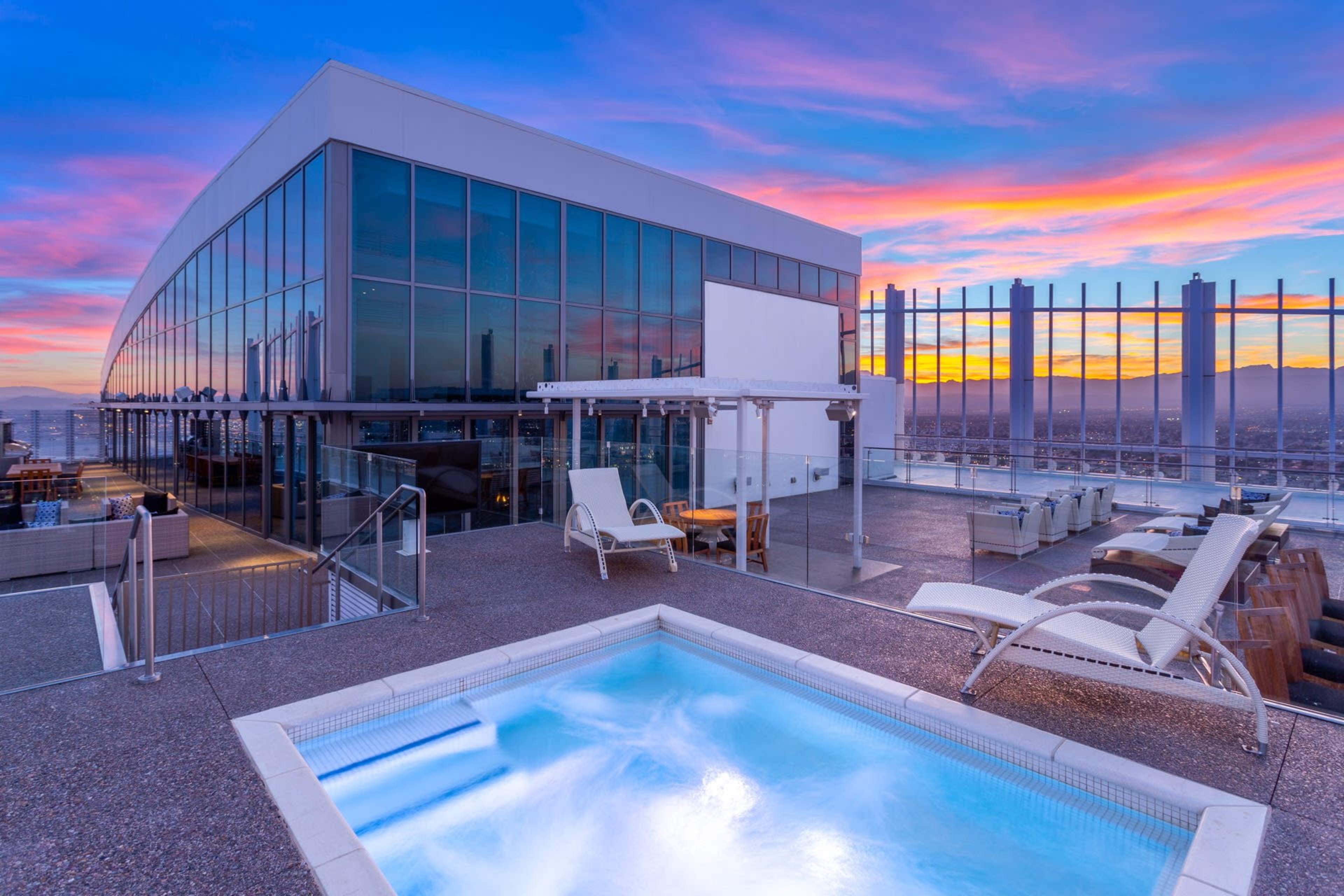 The image shows a modern rooftop terrace featuring a hot tub, lounge chairs, and floor-to-ceiling glass walls with a colorful sunset in the background.