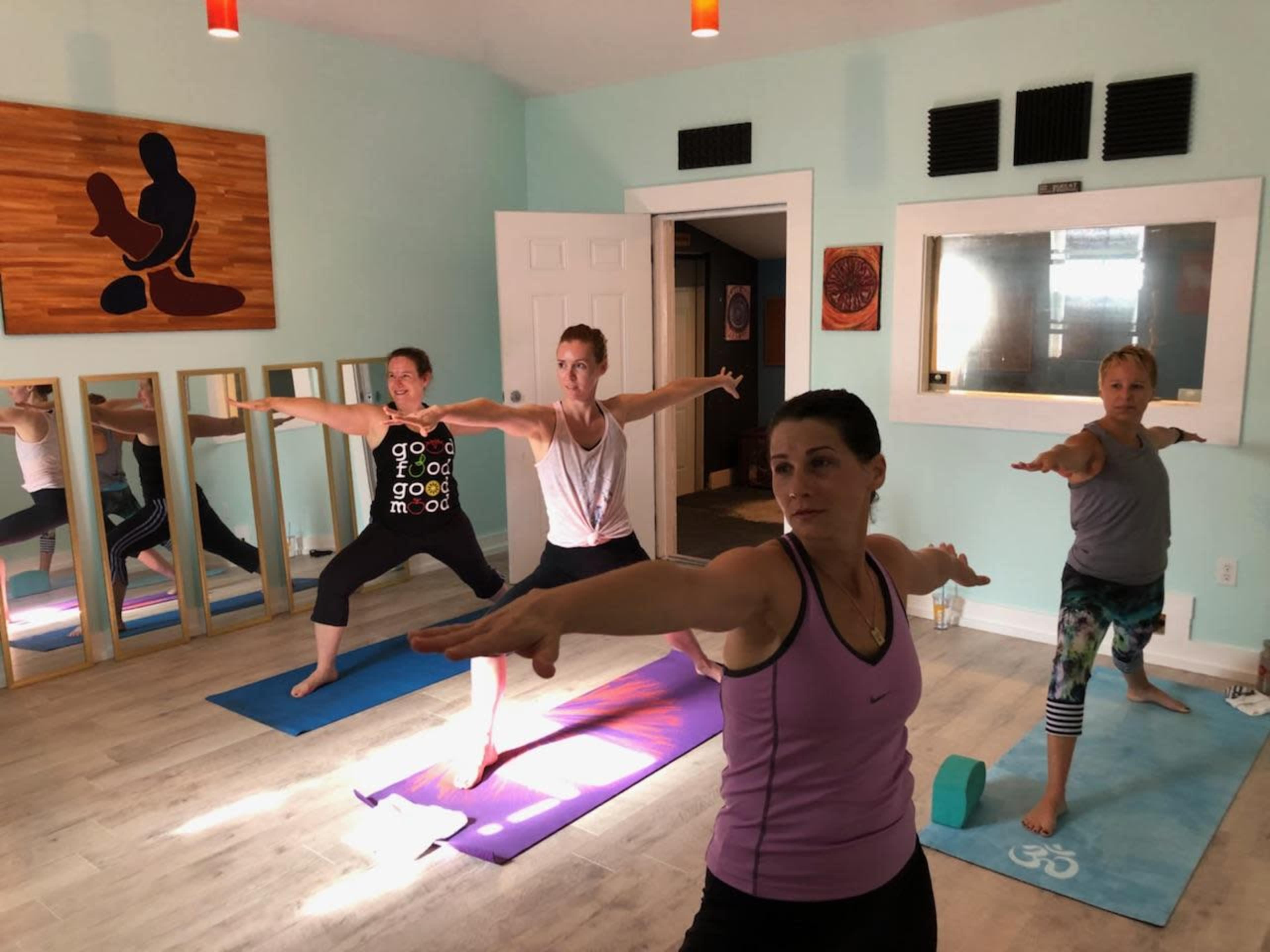 A group of four women practice yoga in a brightly lit studio with mirrors and colorful mats.