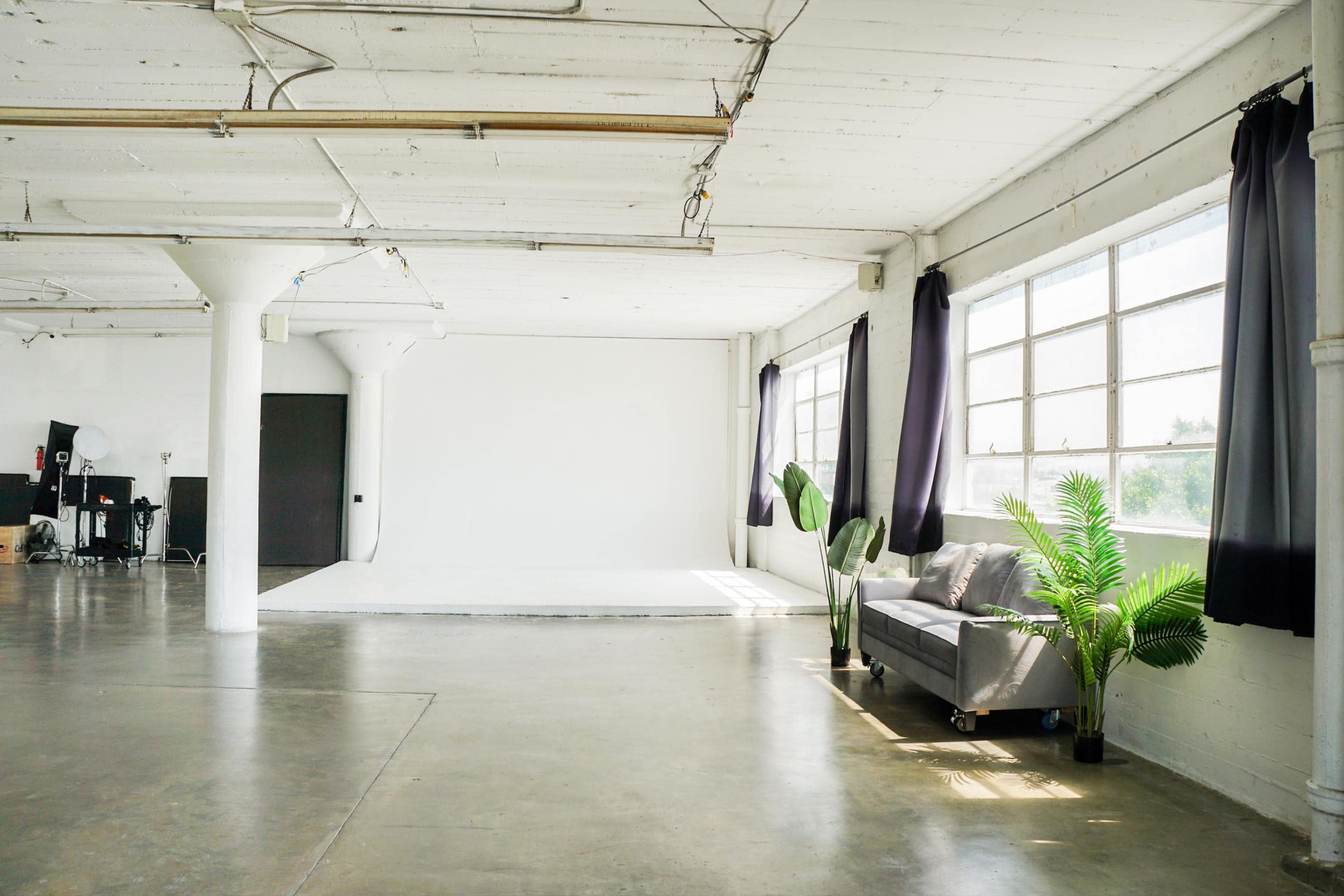 The image shows a spacious, minimalist studio with large windows, a white backdrop, a gray sofa, and a potted plant.