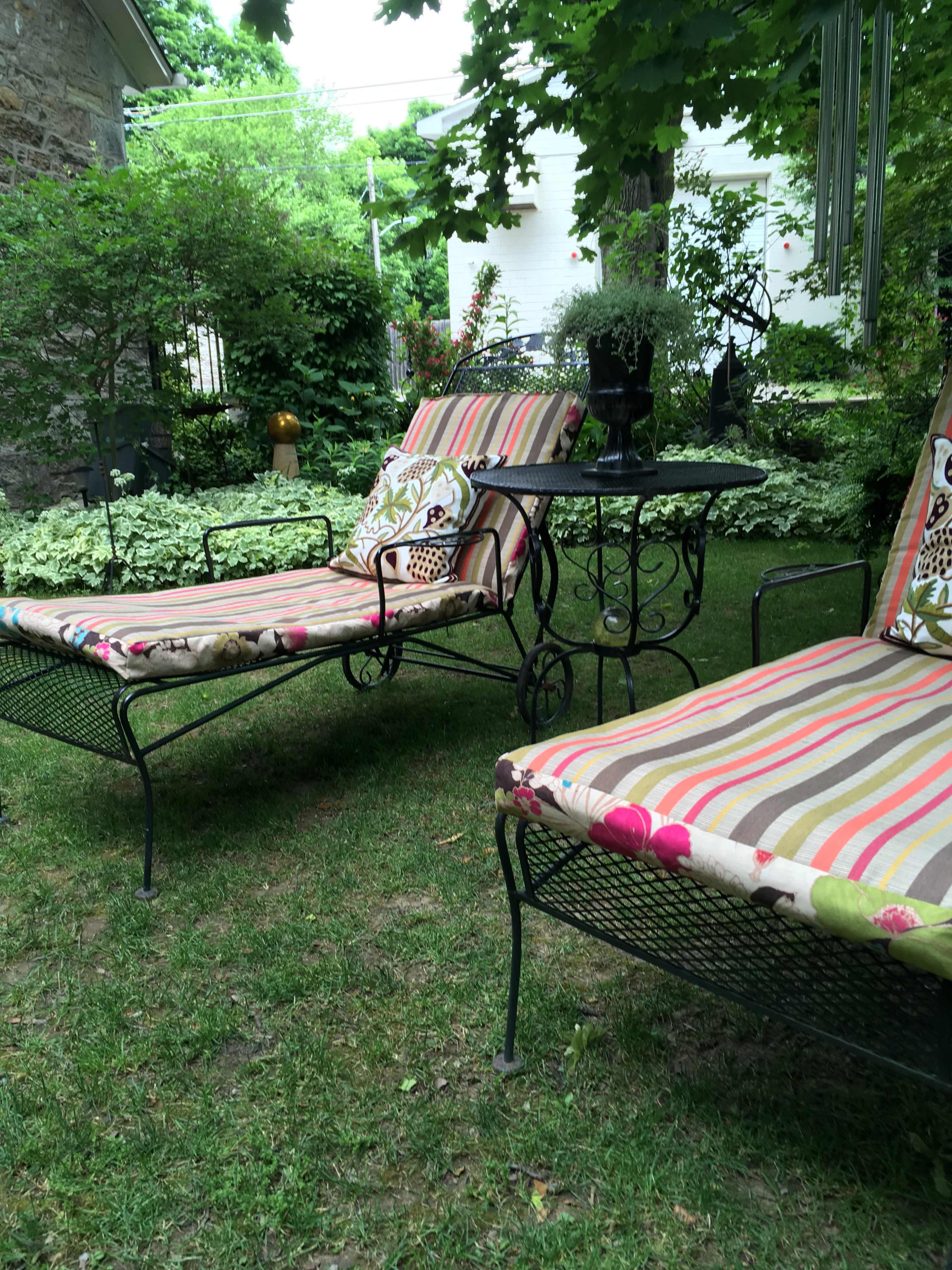 Two striped lounge chairs with decorative cushions sit beside a small metal table in a grassy garden surrounded by greenery.