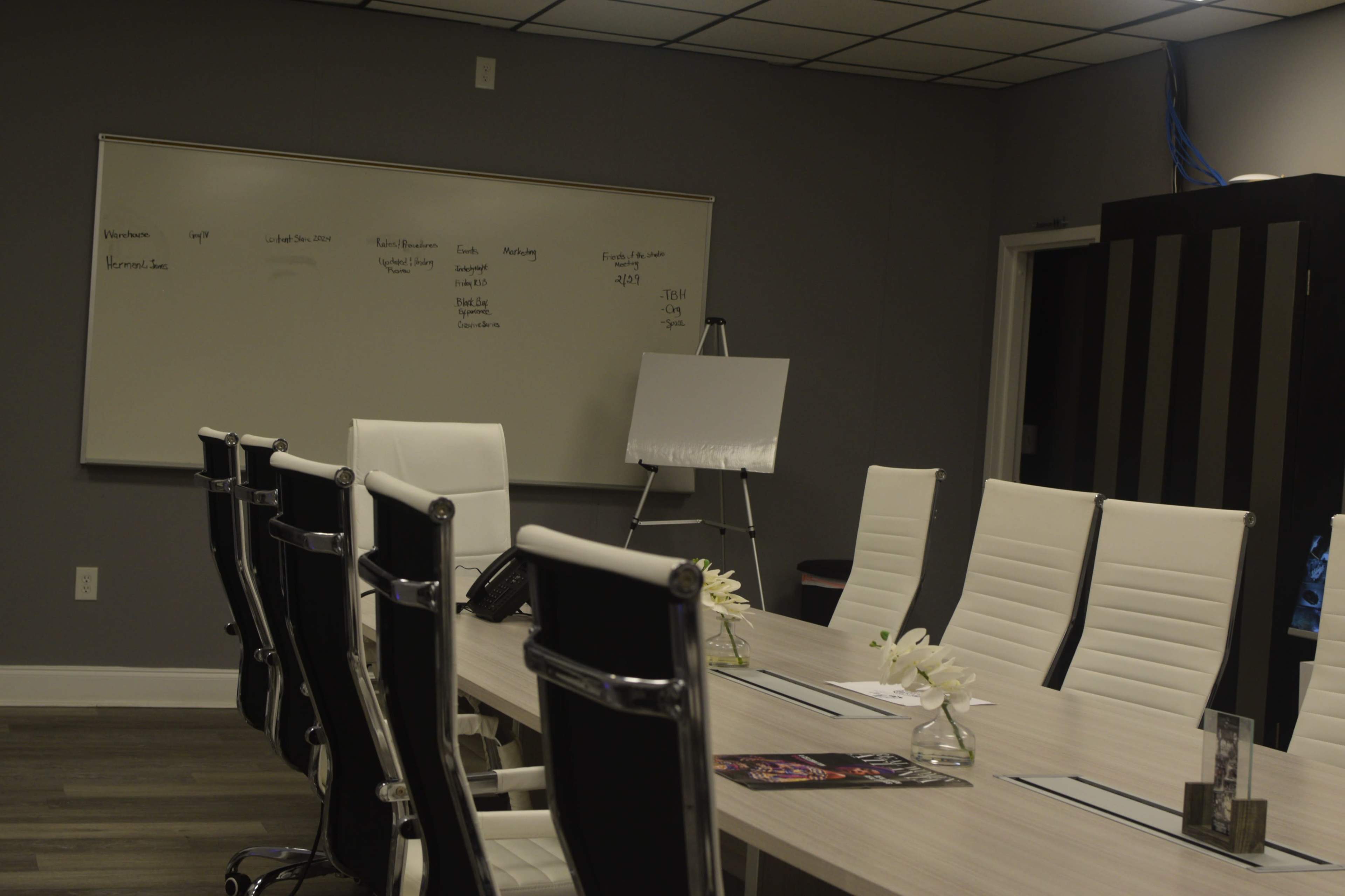 A modern conference room is set up with a long table surrounded by white chairs, a whiteboard on the wall, and an easel with a blank sheet nearby.