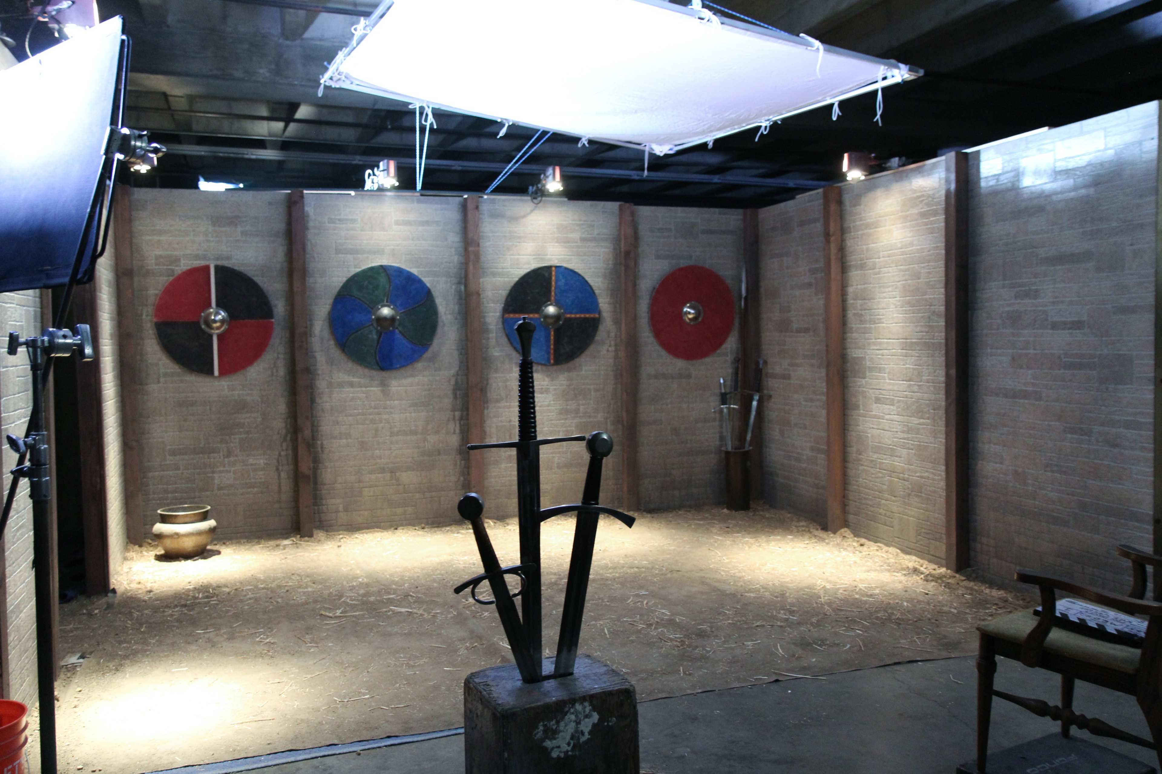 The image shows a sparsely furnished interior resembling a historical setting, featuring three shields on the wall and a large sword displayed on a stone pedestal.