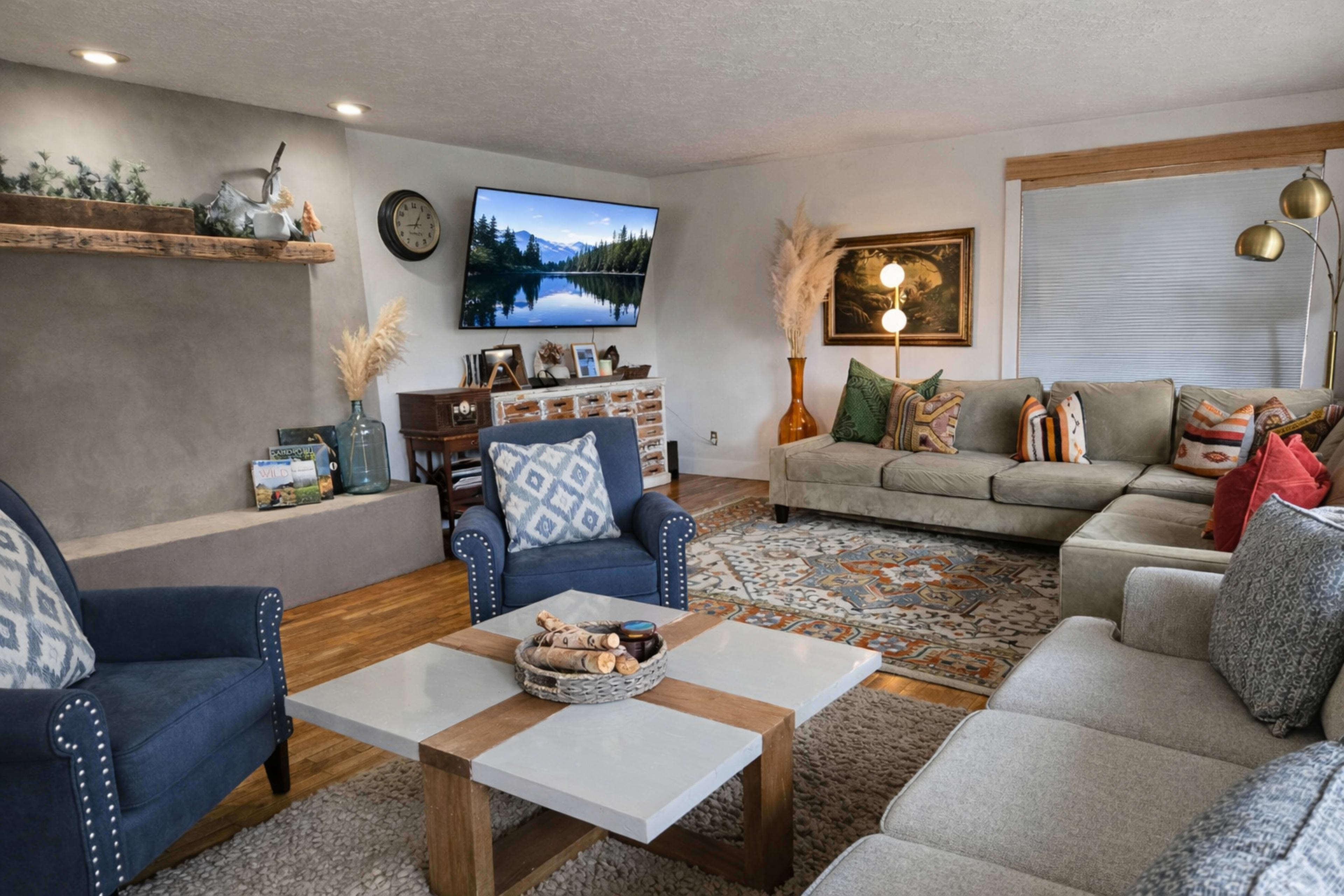 The living room features a comfortable seating arrangement with a large sofa, two blue armchairs, a coffee table, and a mounted television displaying a scenic landscape, alongside decorative elements like plants and framed artwork.
