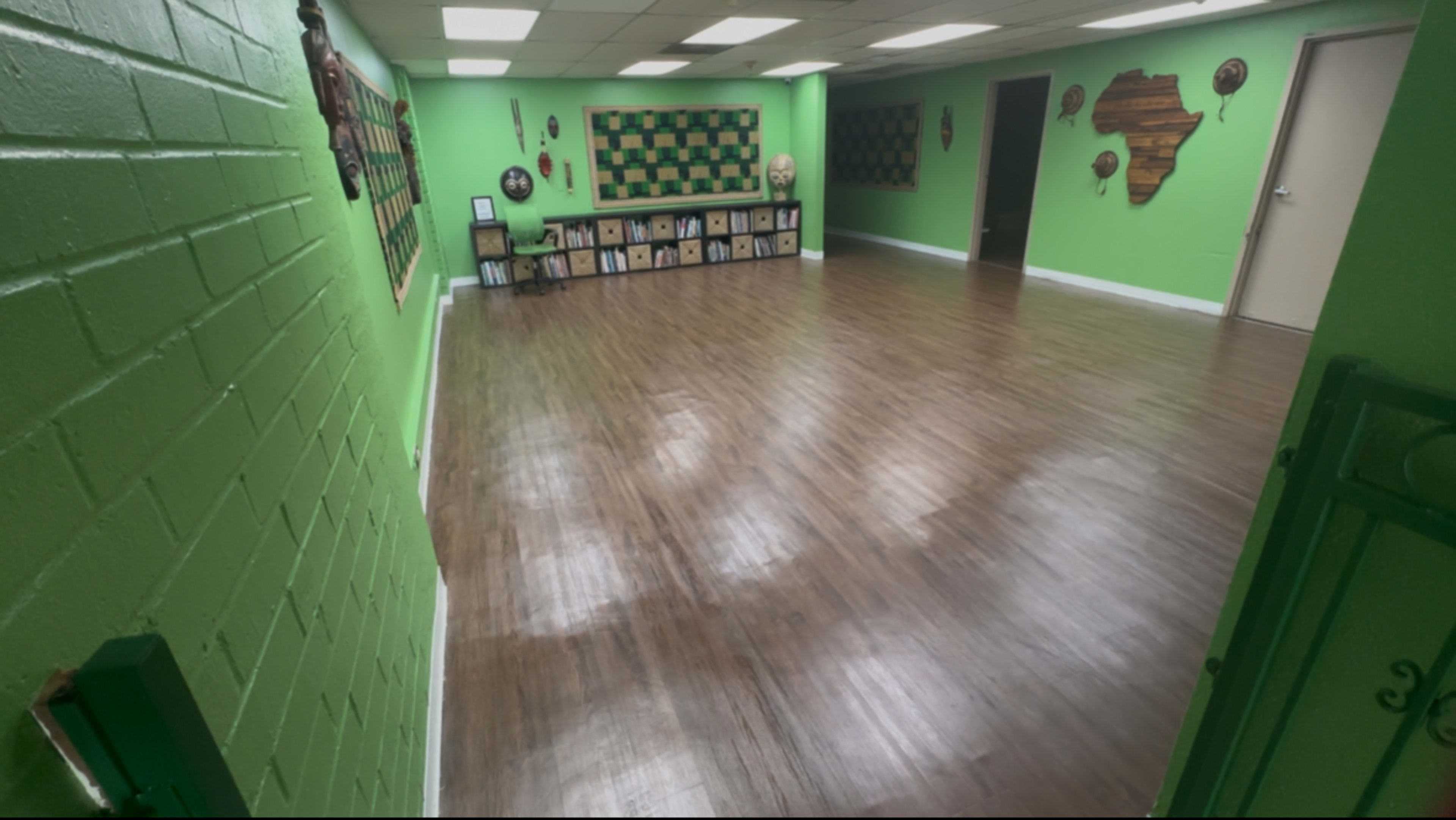 The image shows an empty room with green walls and a wooden floor, featuring decorative wall art and shelves lined with framed items.
