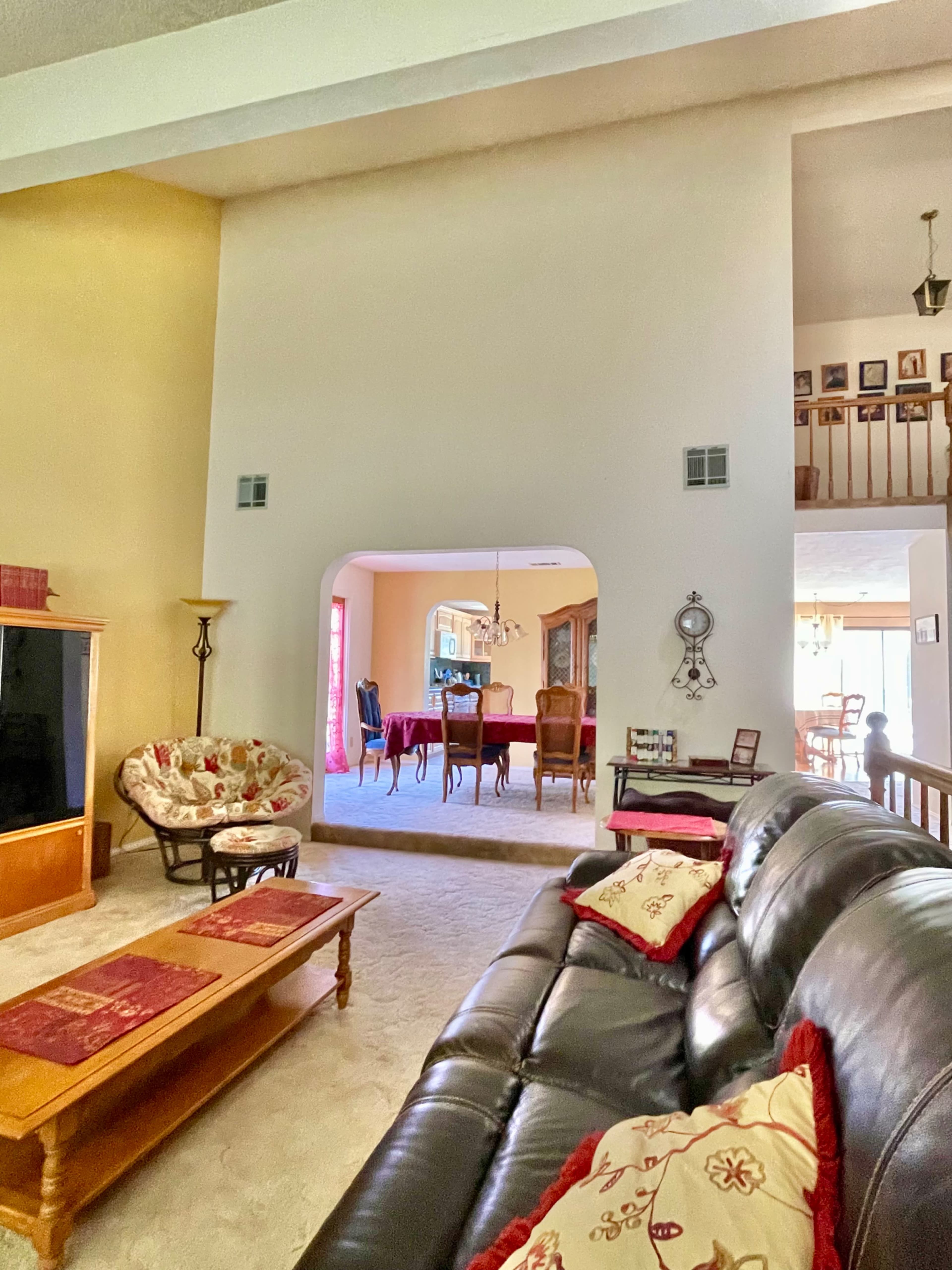 A living room features a large window, a black leather sofa with decorative pillows, and a view of a dining area in the background.