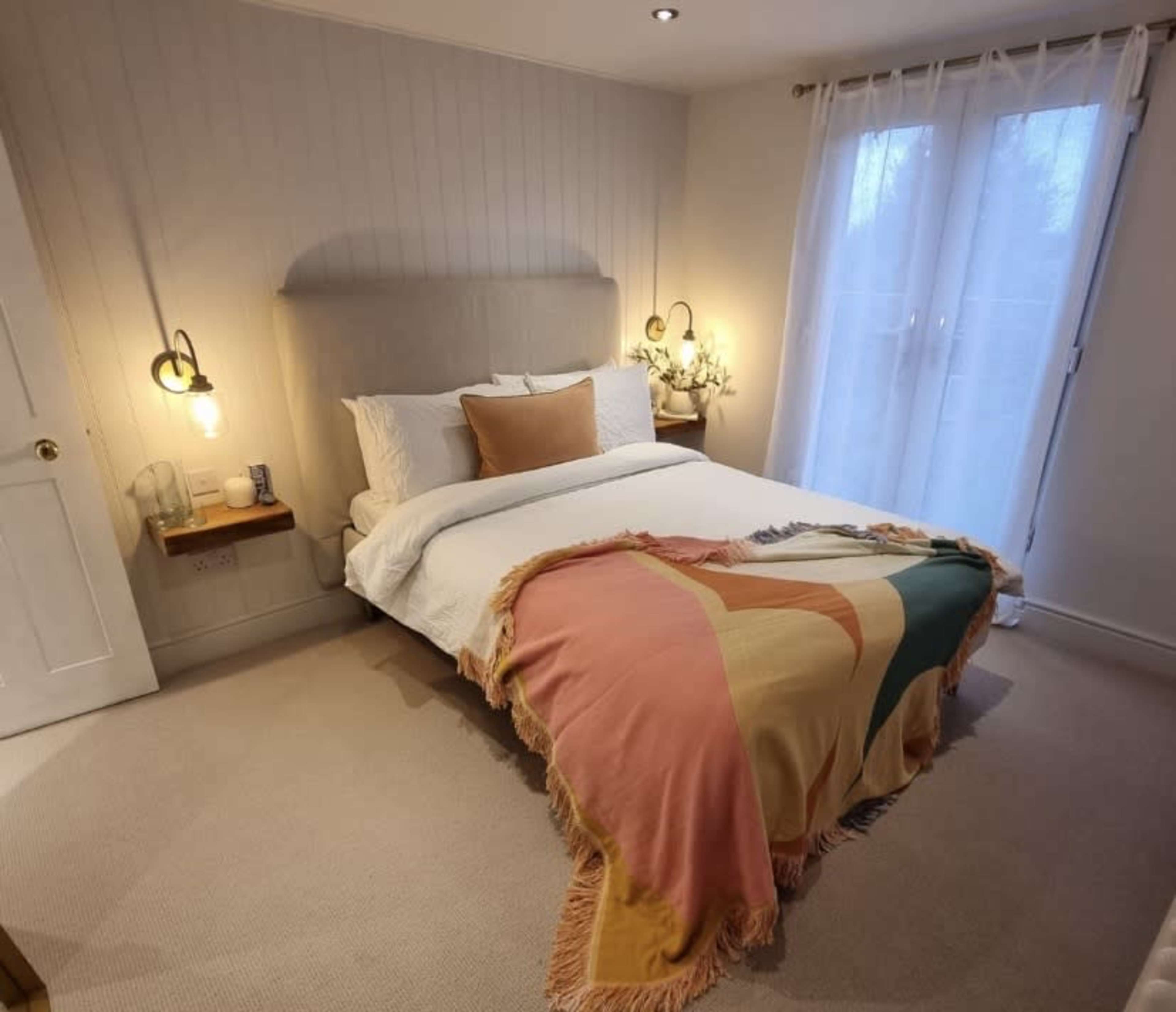 The image shows a well-lit bedroom featuring a neatly made bed with a colorful throw blanket, soft bedding, and sheer curtains letting in natural light.