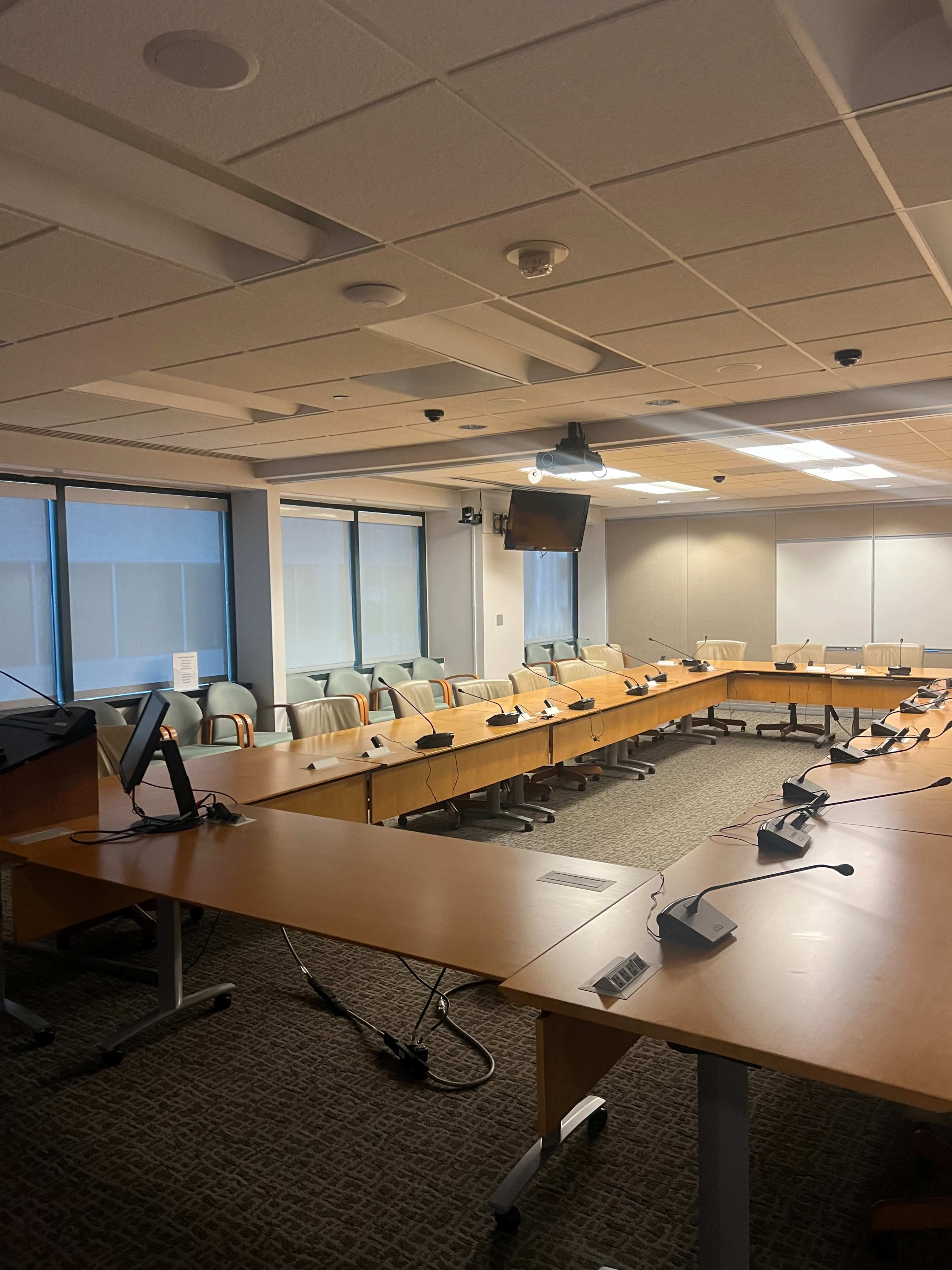 A conference room with a large round table equipped with multiple microphones and a projection screen.