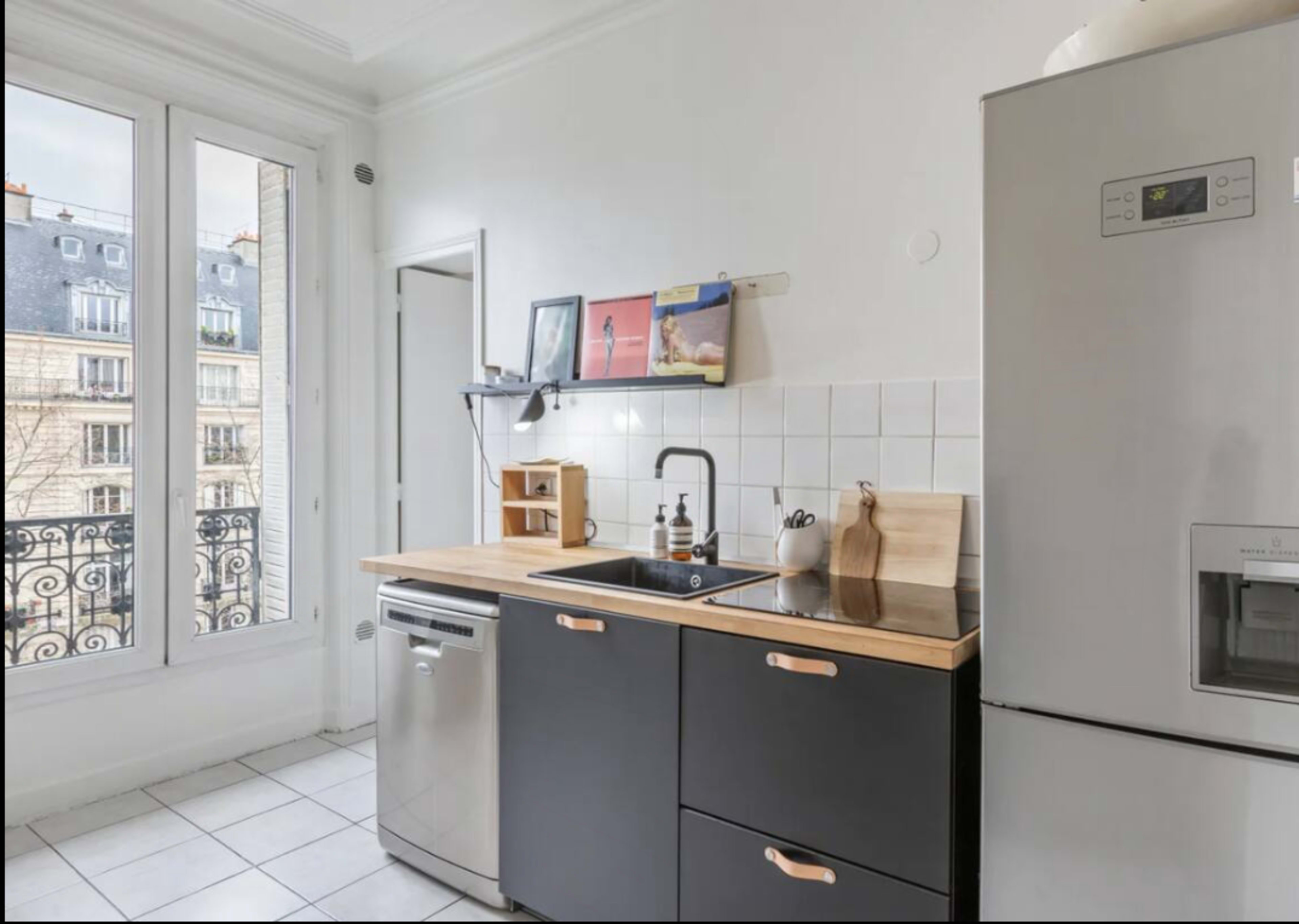 A small kitchen features a stainless steel refrigerator, a wooden countertop with a sink, and a window overlooking a balcony.