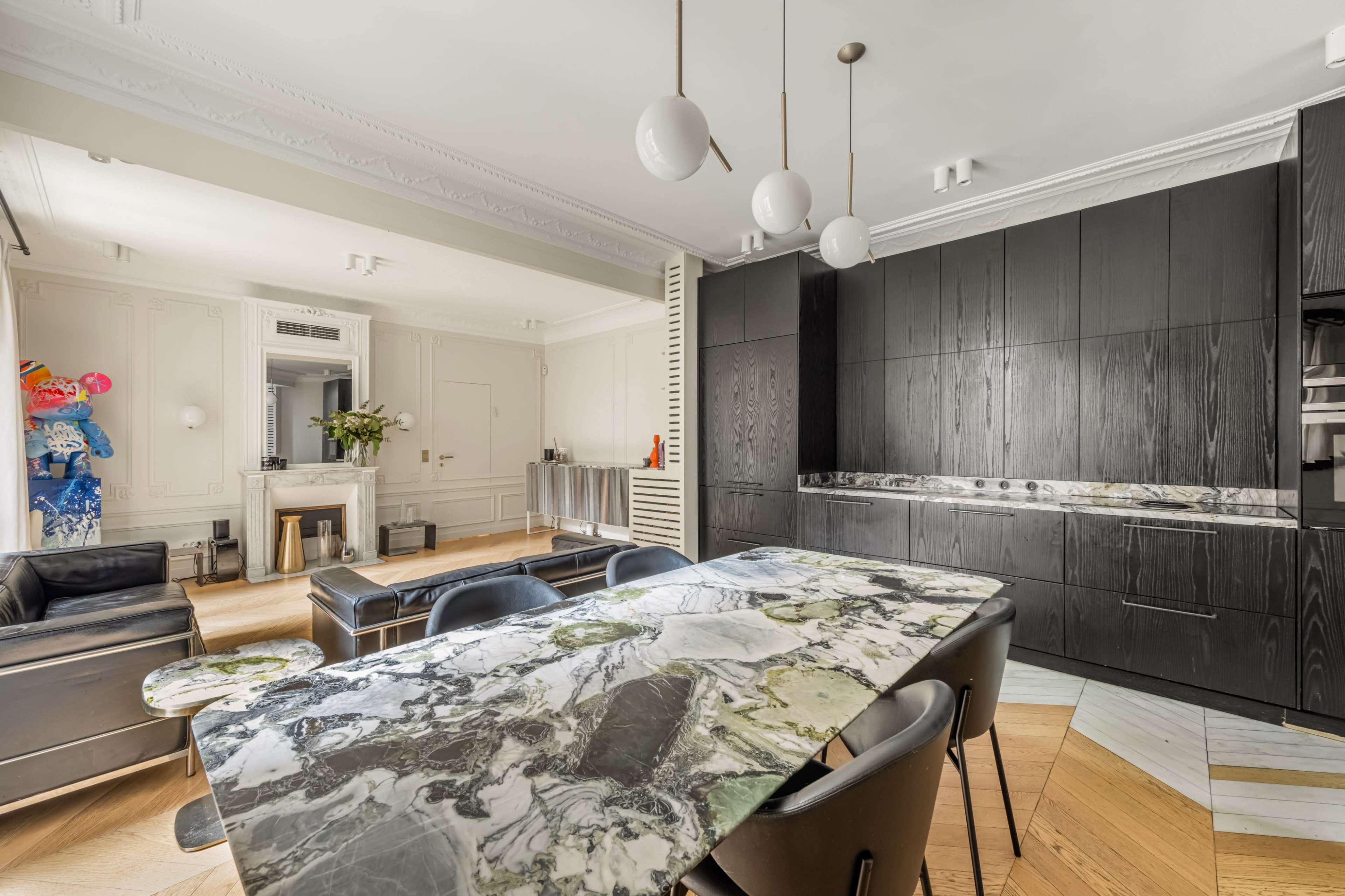 A modern kitchen features a marble dining table surrounded by black chairs, with sleek dark cabinetry and a bright living area in the background.