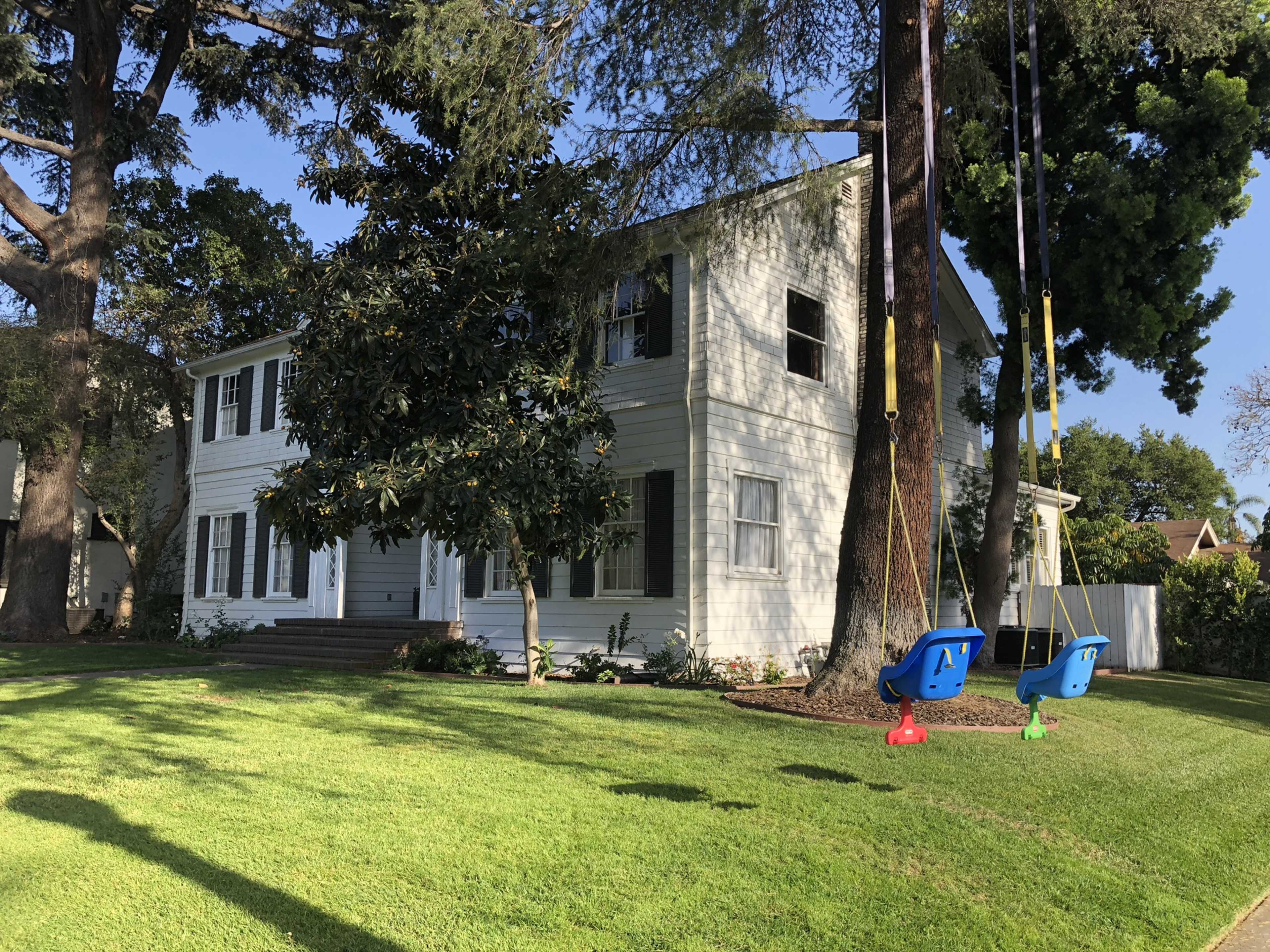 A two-story white house with green shutters sits on a well-maintained lawn, featuring two swings hanging from tree branches nearby.