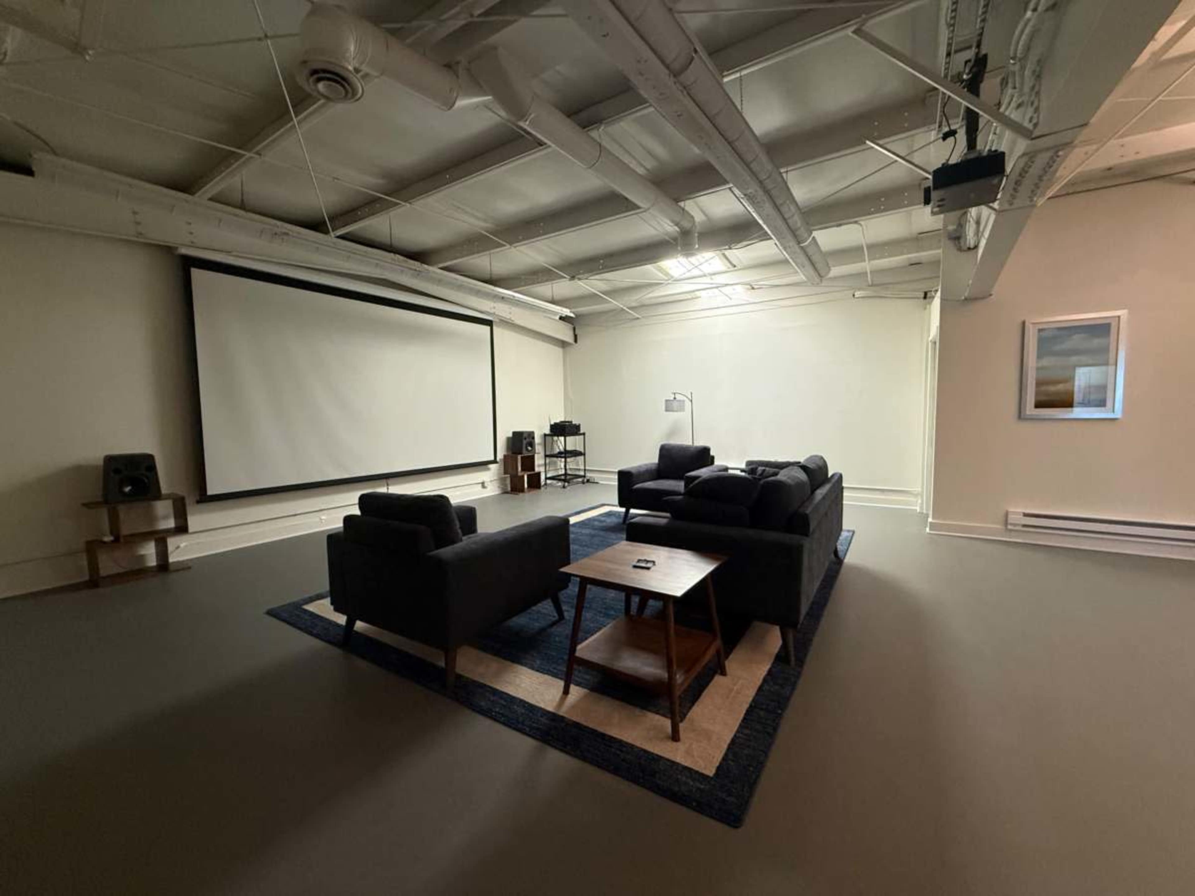 The image shows a spacious room with a large projection screen at one end, furnished with a dark sofa set and a coffee table on a light rug.