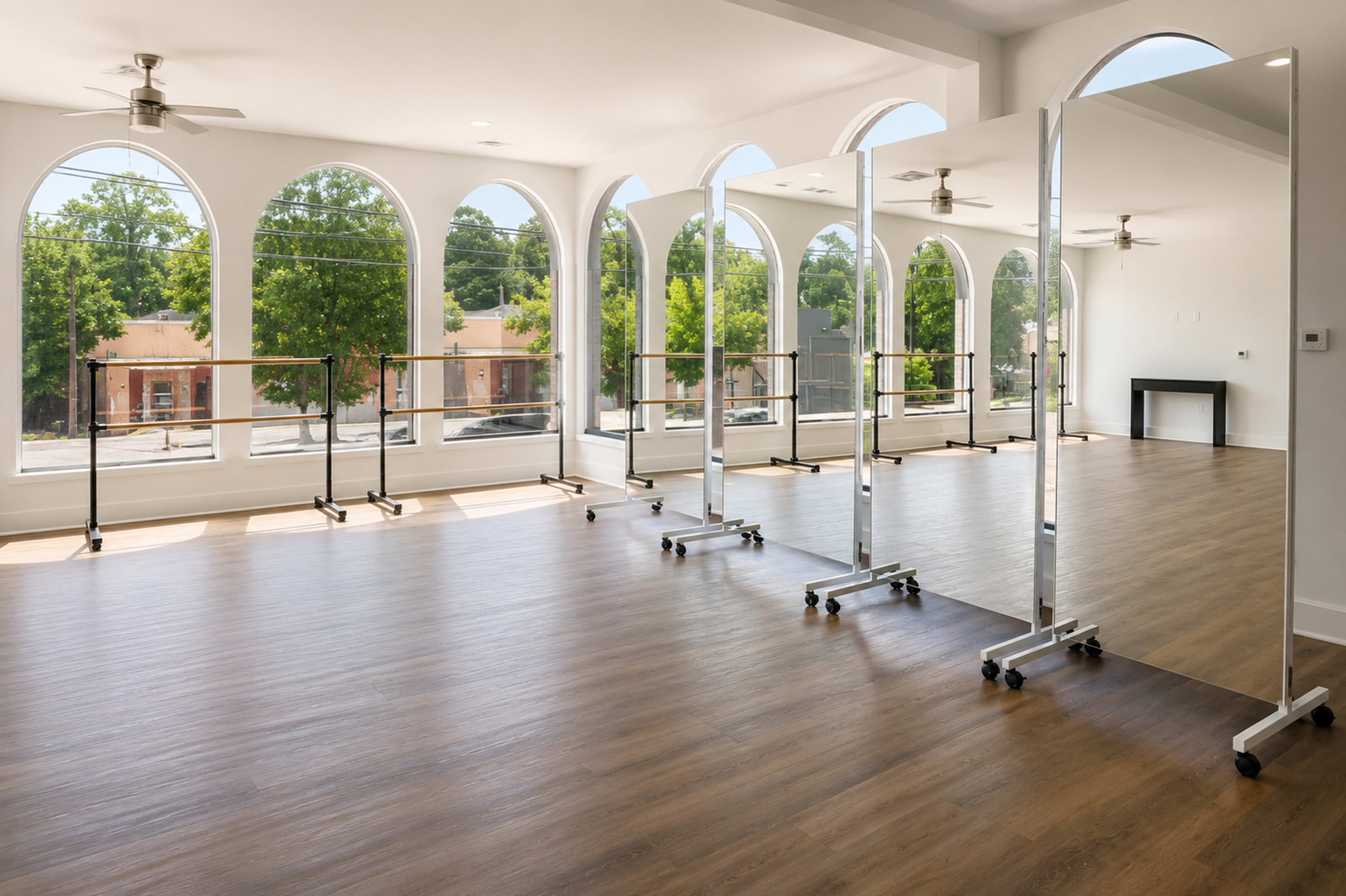The image shows a spacious dance studio with large arched windows, wooden flooring, and several movable mirrors arranged along one side.