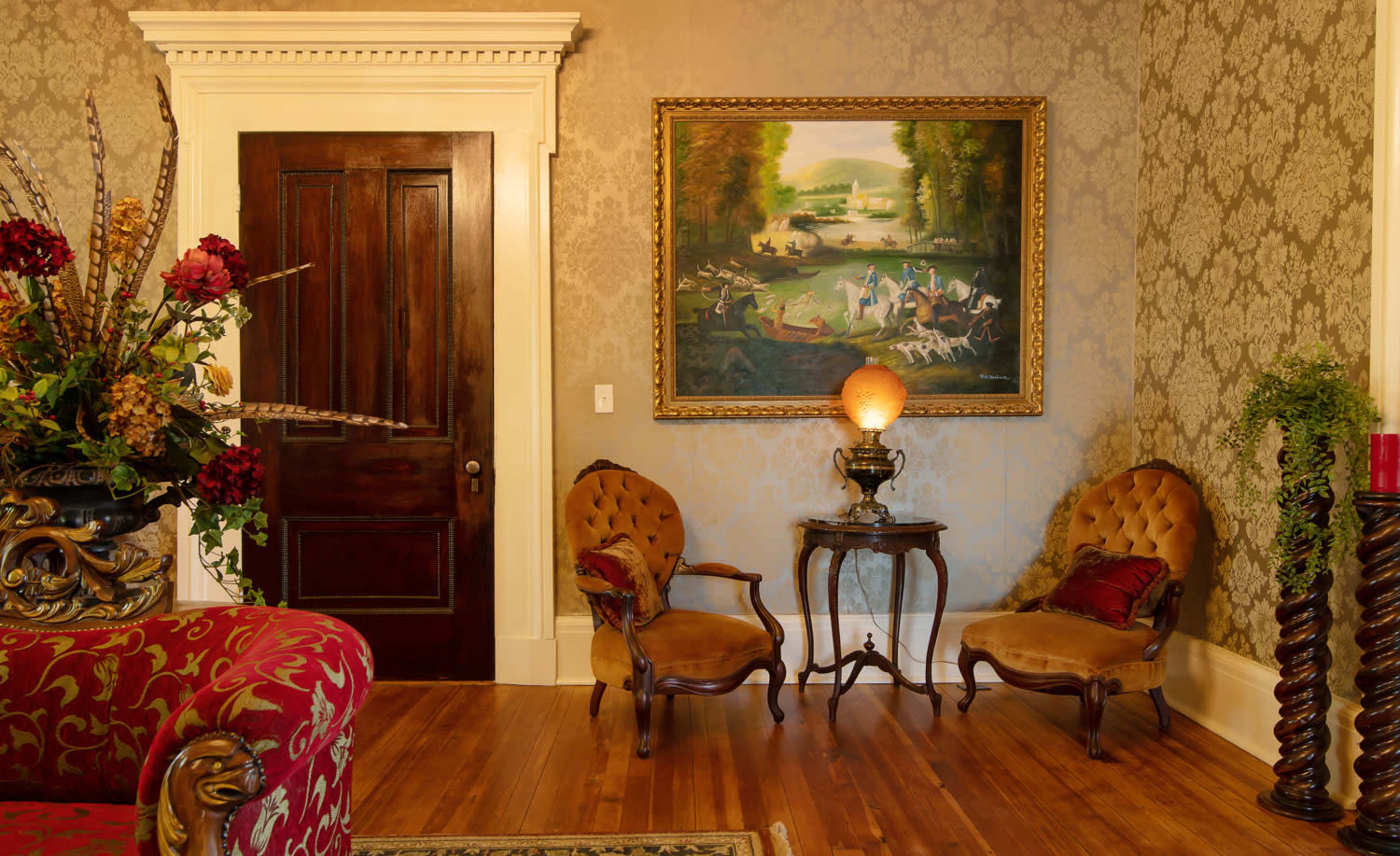 A parlor with two upholstered chairs, a small table, a large painting on the wall, and a decorative flower arrangement next to the entrance door.