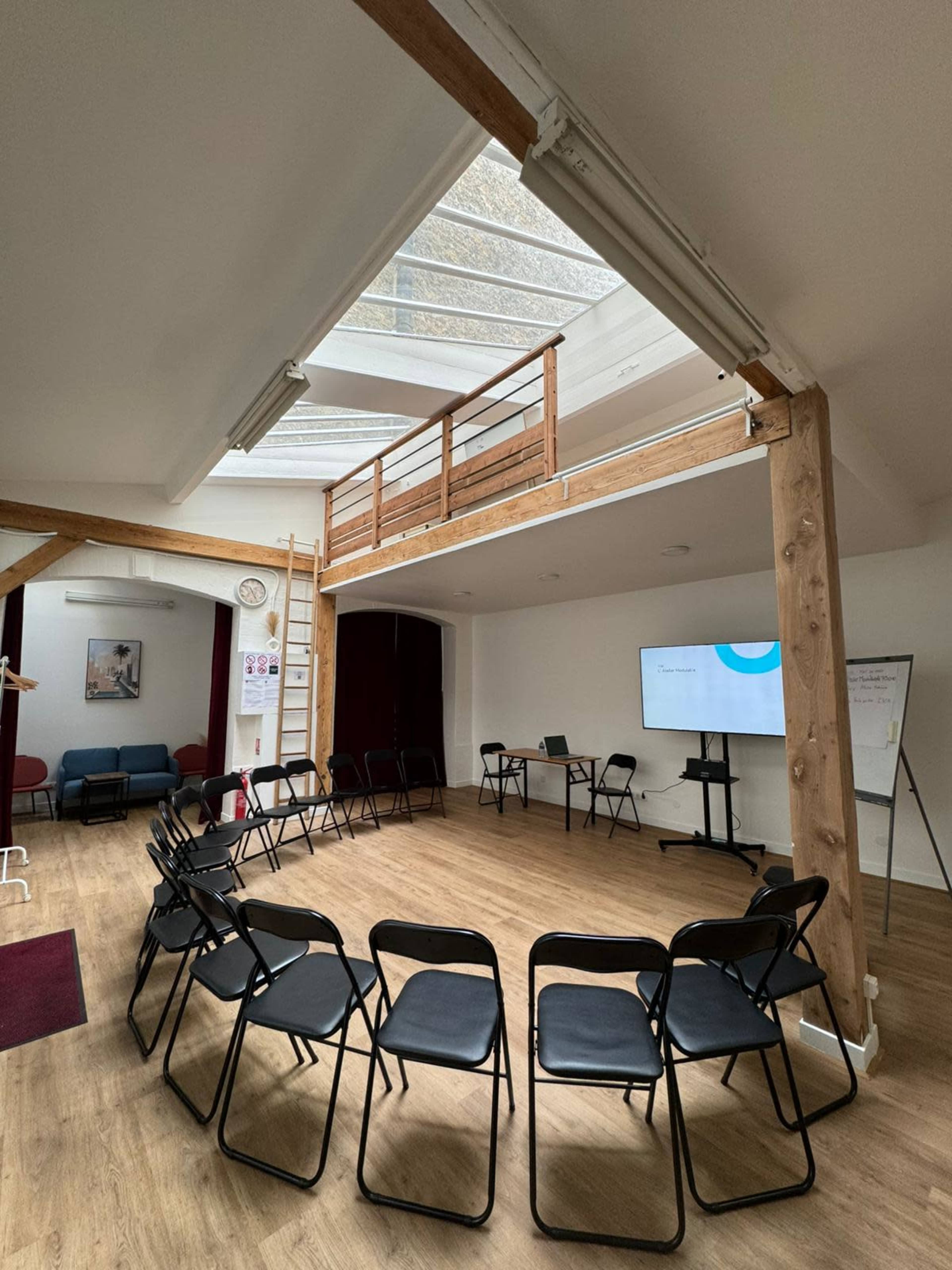 The image shows a spacious meeting room with a circular arrangement of black chairs, a wooden balcony overhead, and a screen displaying a presentation.