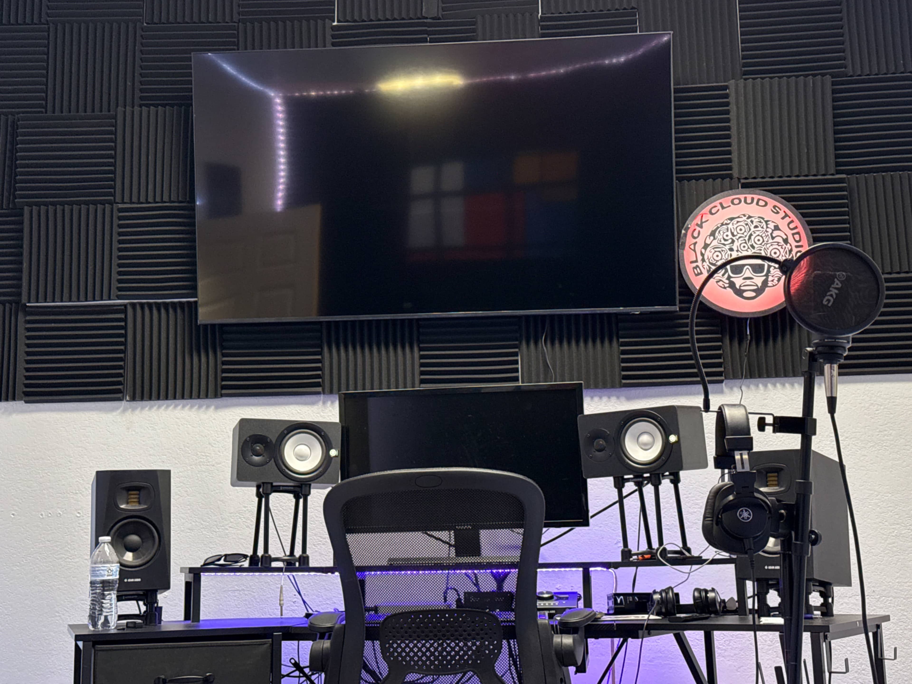 The image shows a music studio setup with a large wall-mounted TV above a black desk featuring audio equipment, monitors, and a microphone.