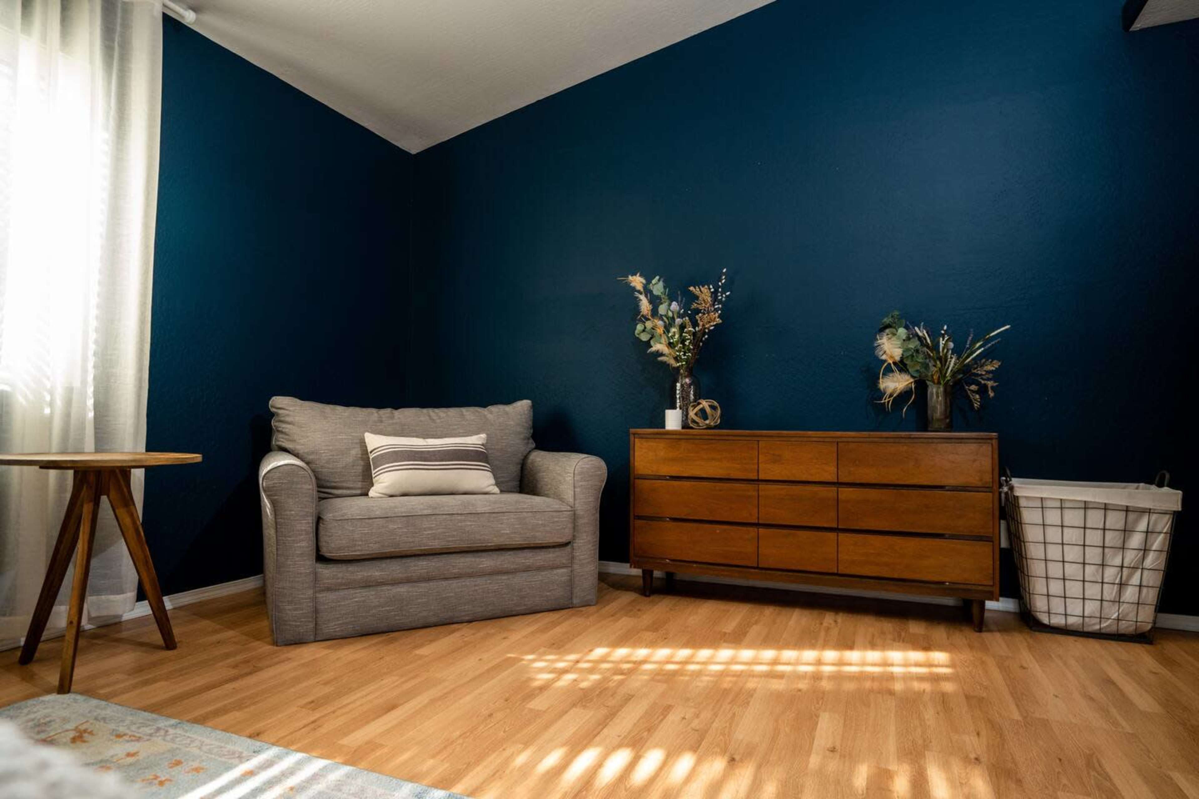 The image shows a room with a gray armchair, a wooden dresser, a decorative basket, and floral arrangements on display against a dark blue wall.