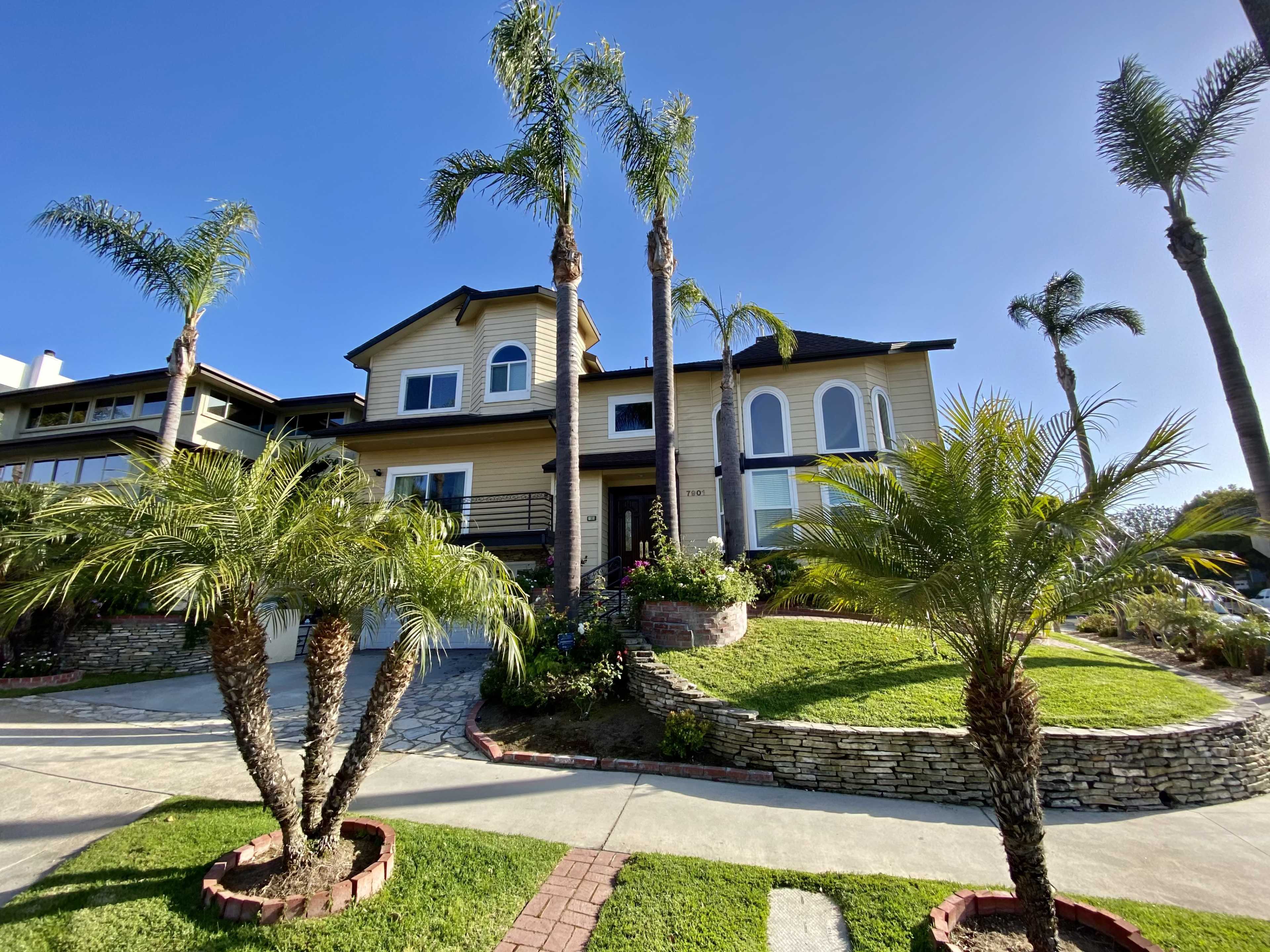 A two-story house with a large front yard and palm trees is situated on a paved driveway.