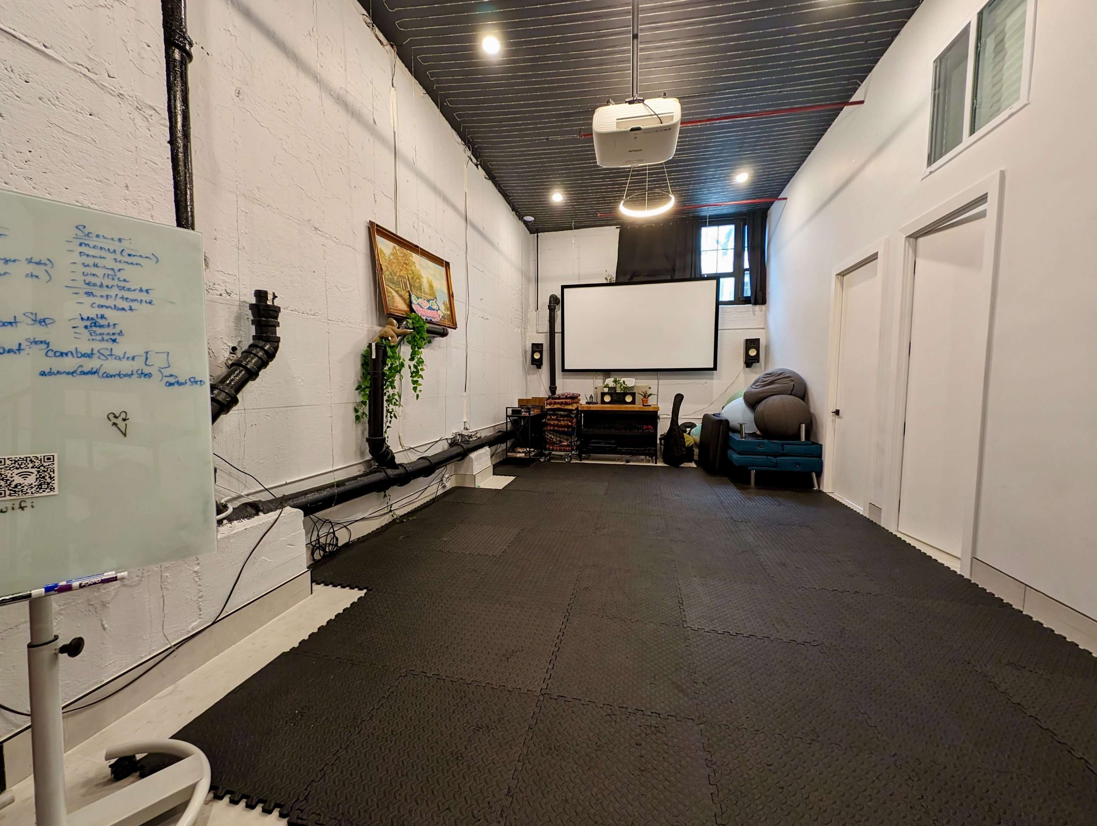 An empty room with black flooring, a projector mounted on the ceiling, a large screen at the front, and a whiteboard on the left wall.