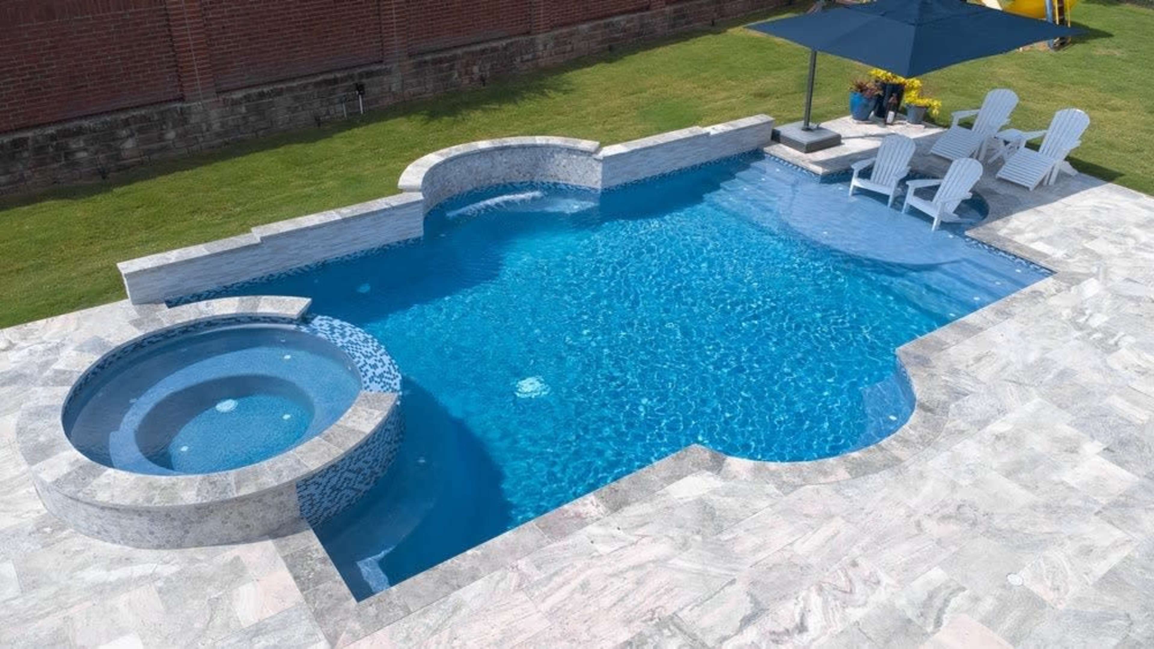 The image shows a blue swimming pool with a connected hot tub, surrounded by a patio and several white lounge chairs under an umbrella.