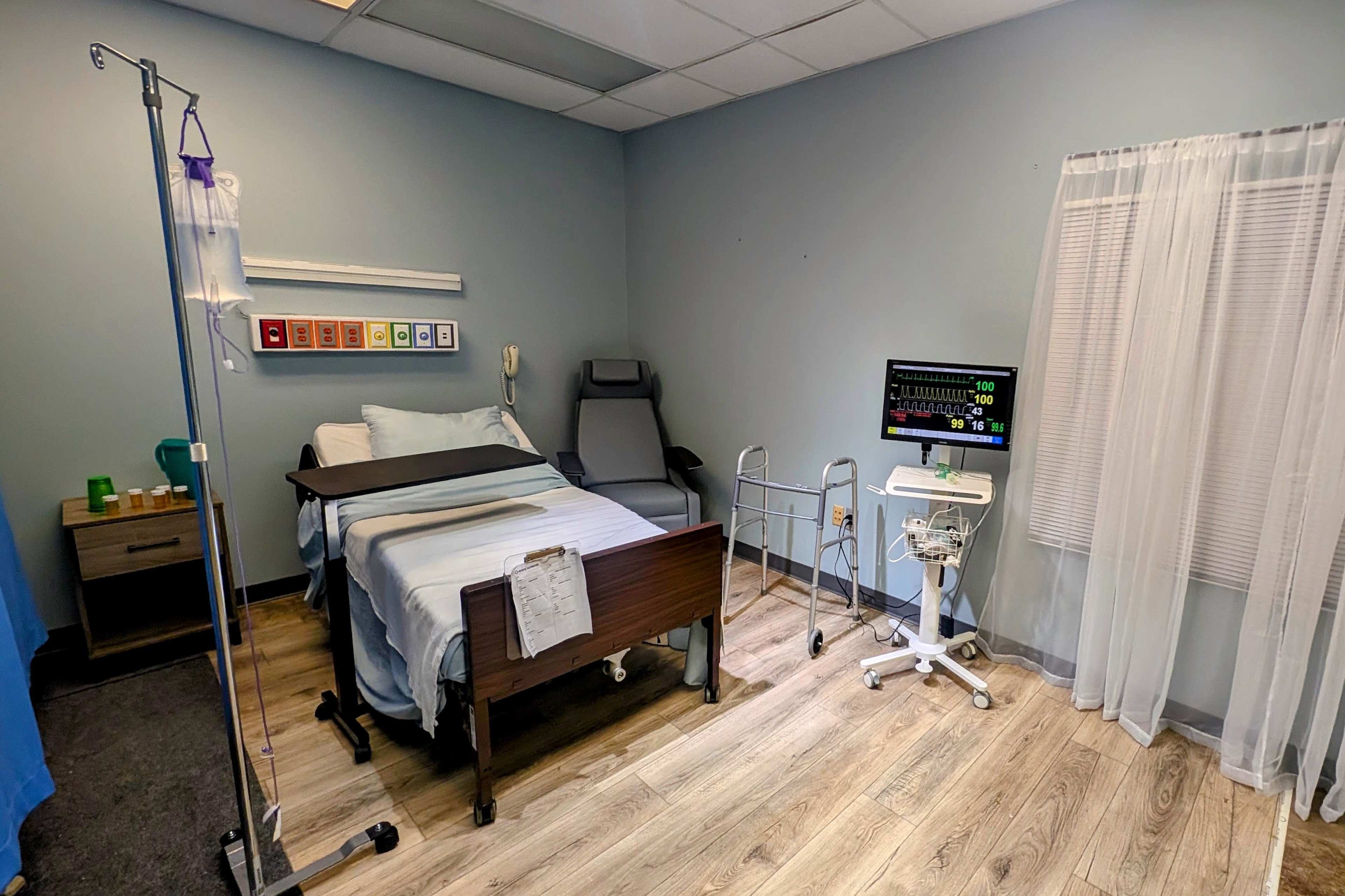 The image shows a hospital room featuring a hospital bed, a reclining chair, a medical monitor, and a privacy curtain.