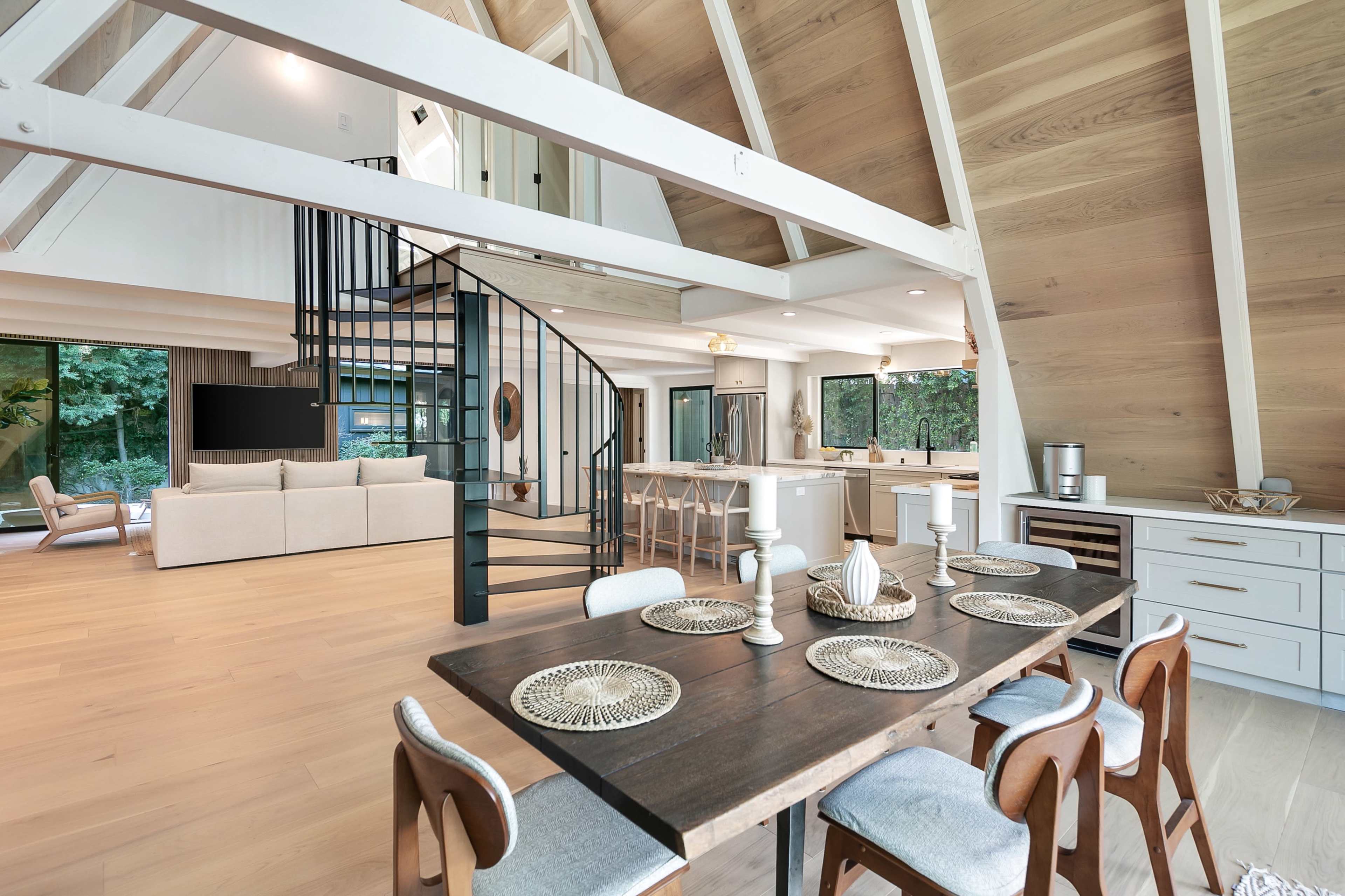 The image shows a modern open-concept living space with a dining area featuring a wooden table set for six, a spiral staircase leading to a loft, and large windows allowing natural light to fill the room.