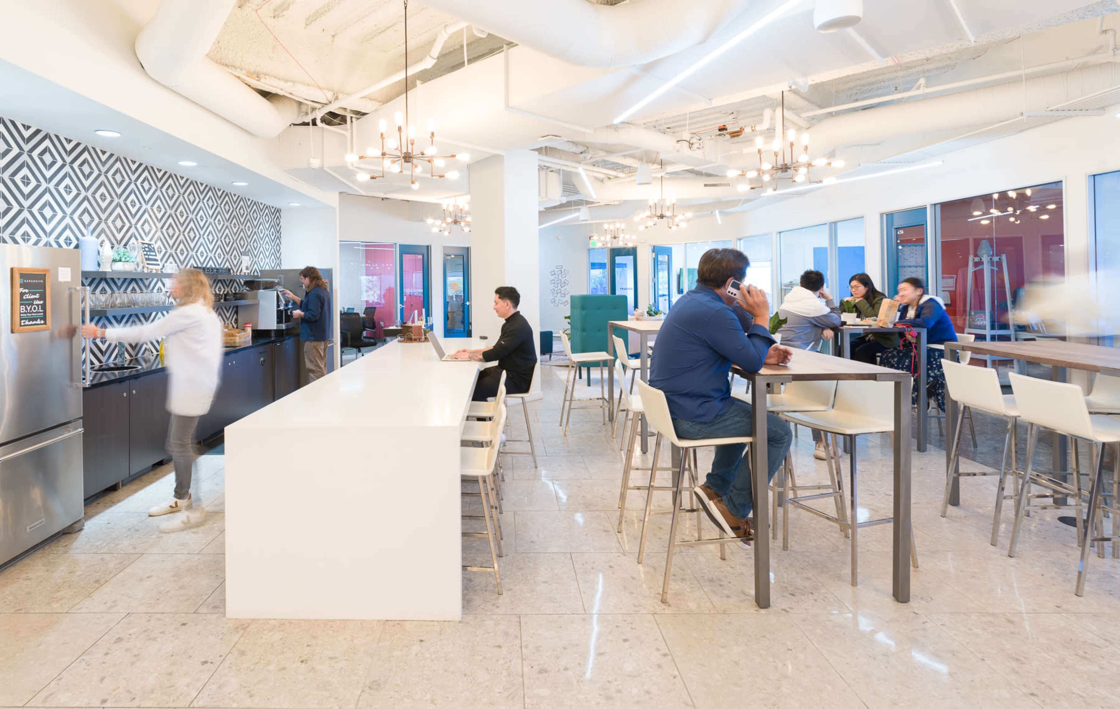 The image shows a modern office kitchen and break area with a long white table, high stools, and a patterned wall, where people are engaged in conversation and preparing food.