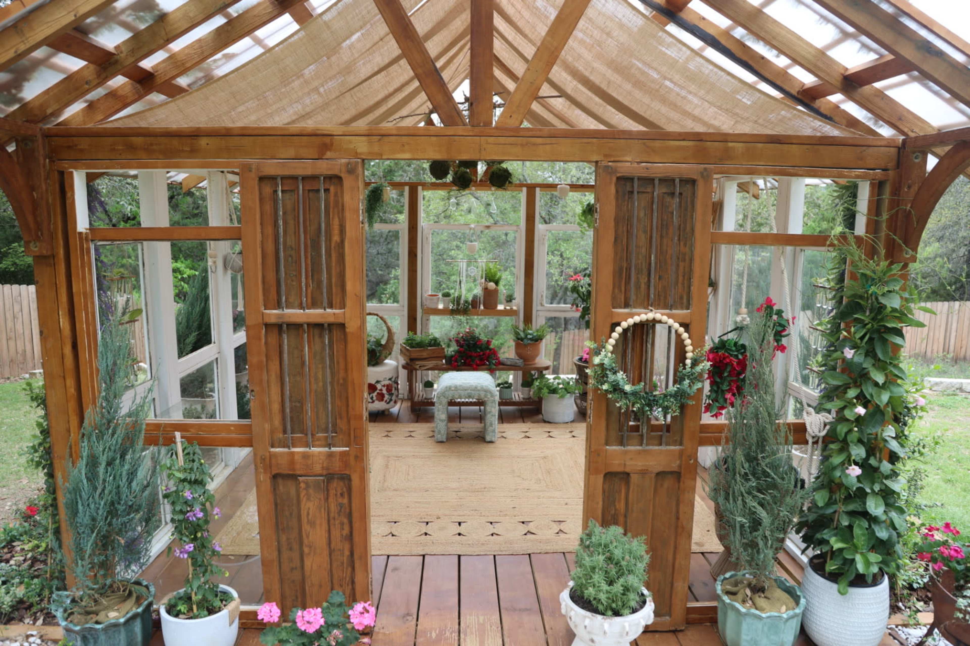The image shows a wooden greenhouse with open doors, featuring various potted plants and a central seating area surrounded by greenery.