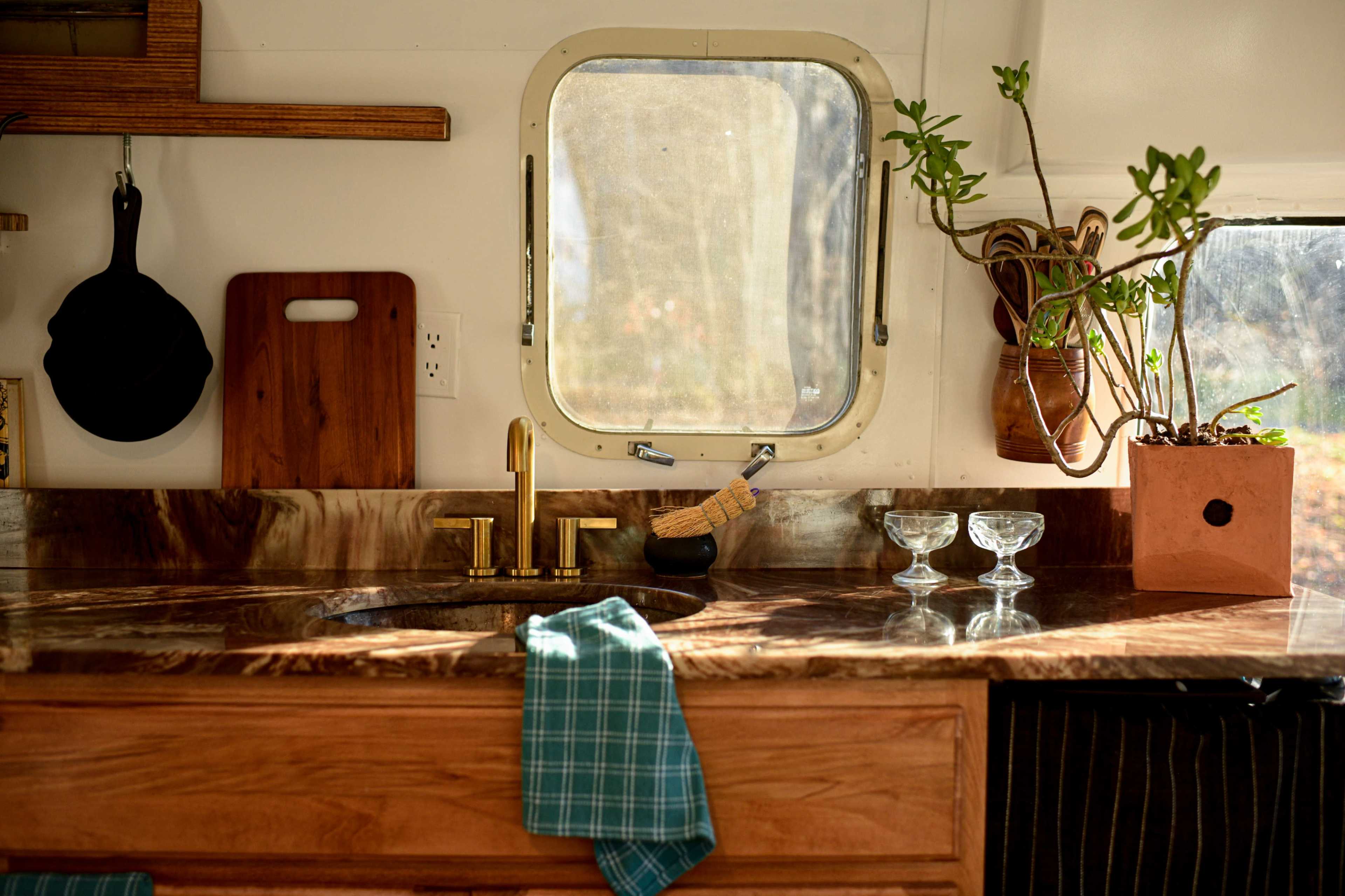 A kitchen countertop with a sink, gold faucet, wooden cutting board, and potted plant near a window.