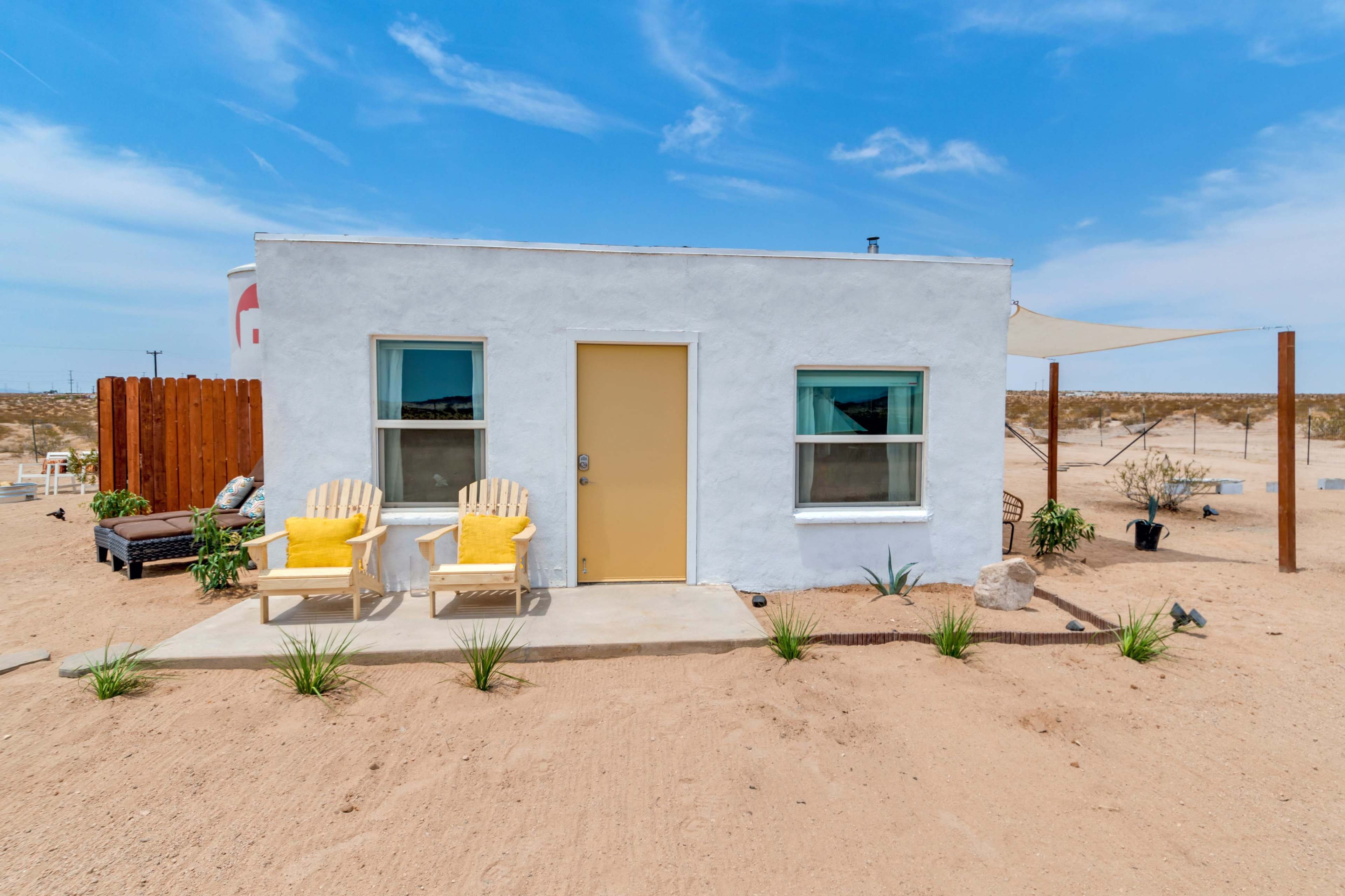 A small white house with a yellow door and two yellow chairs is surrounded by sandy terrain and sparse vegetation.