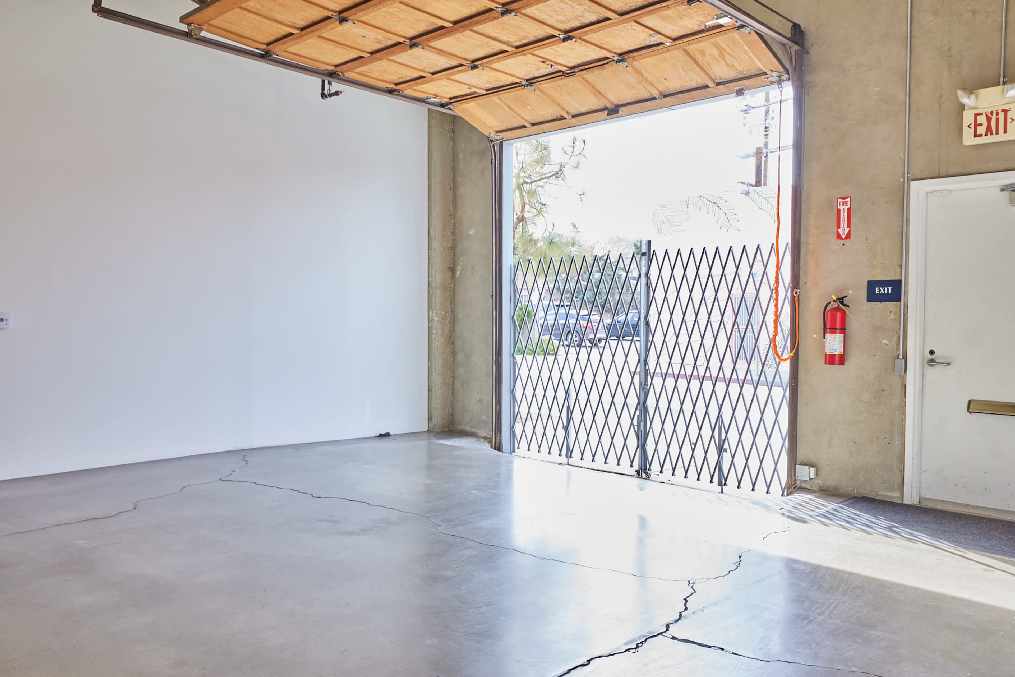 A spacious garage with an open door revealing an exit and a gated entrance, featuring a concrete floor with visible cracks.