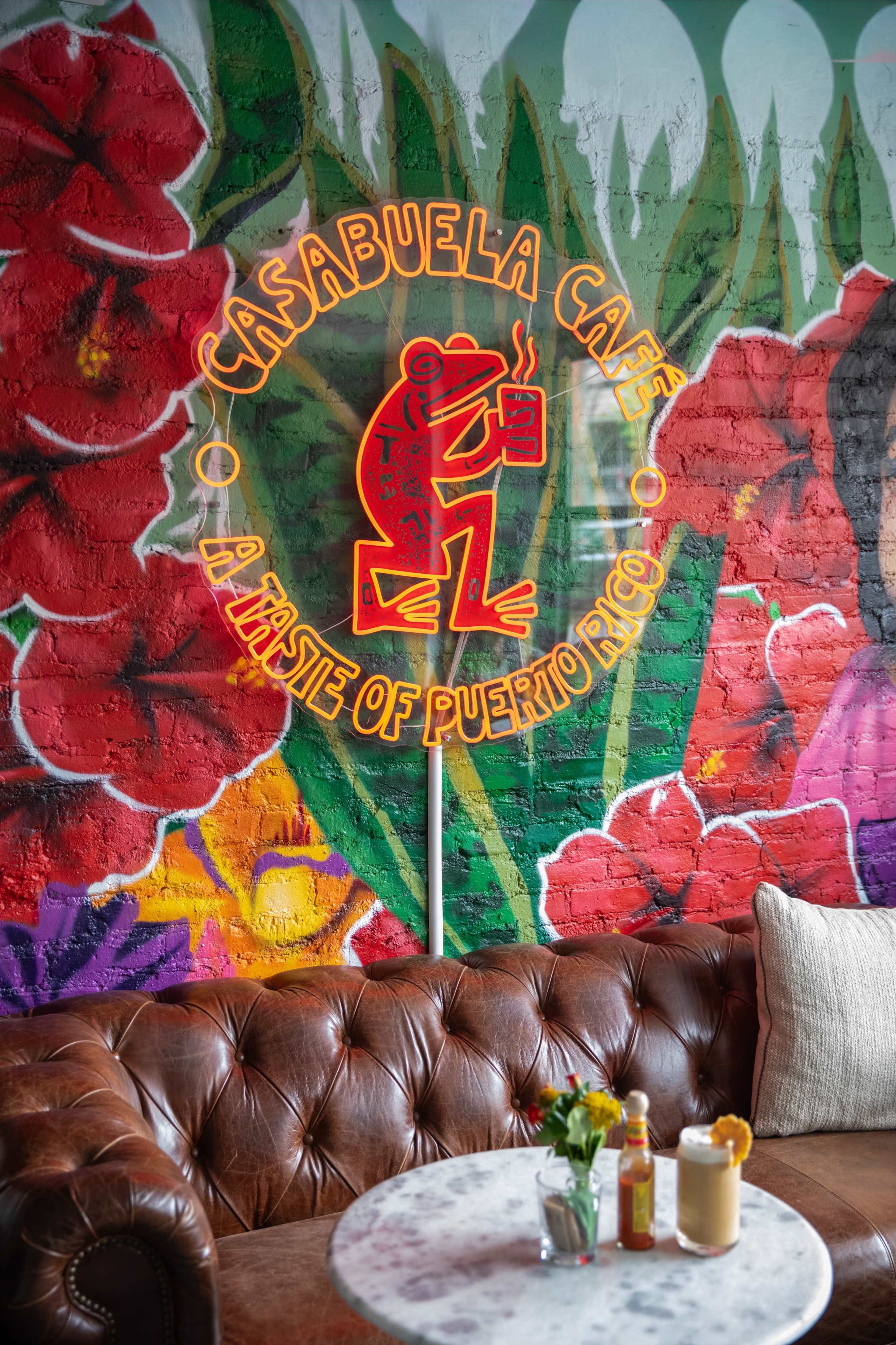 The interior of Casa Abuela Cafe features a vibrant mural with tropical plants and flowers, alongside a neon sign and a brown leather couch with a small table holding drinks.